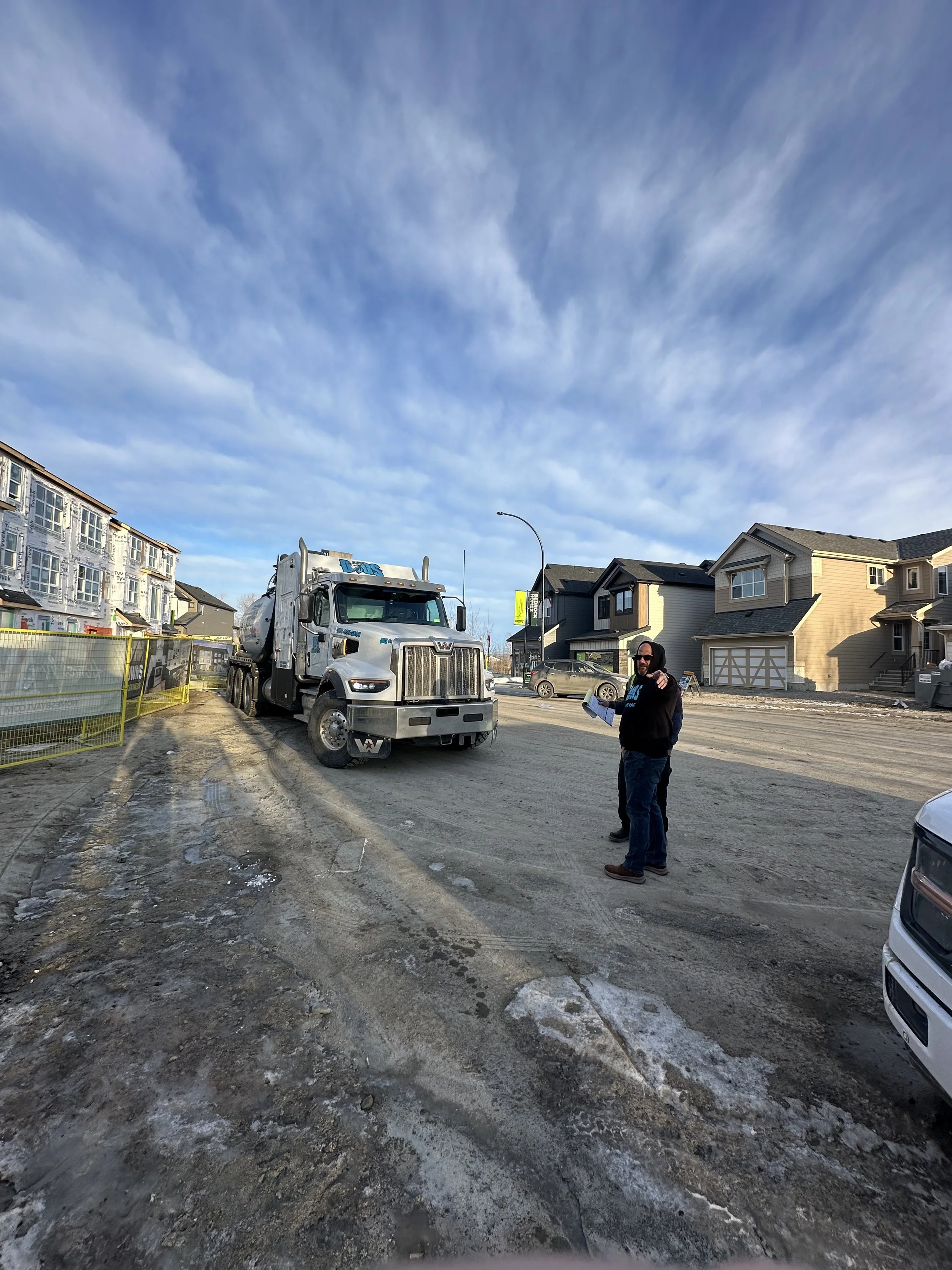 A construction site on a residential street with a large cement truck and two workers, one holding documents, with partially built houses in the background under a partly cloudy sky.
