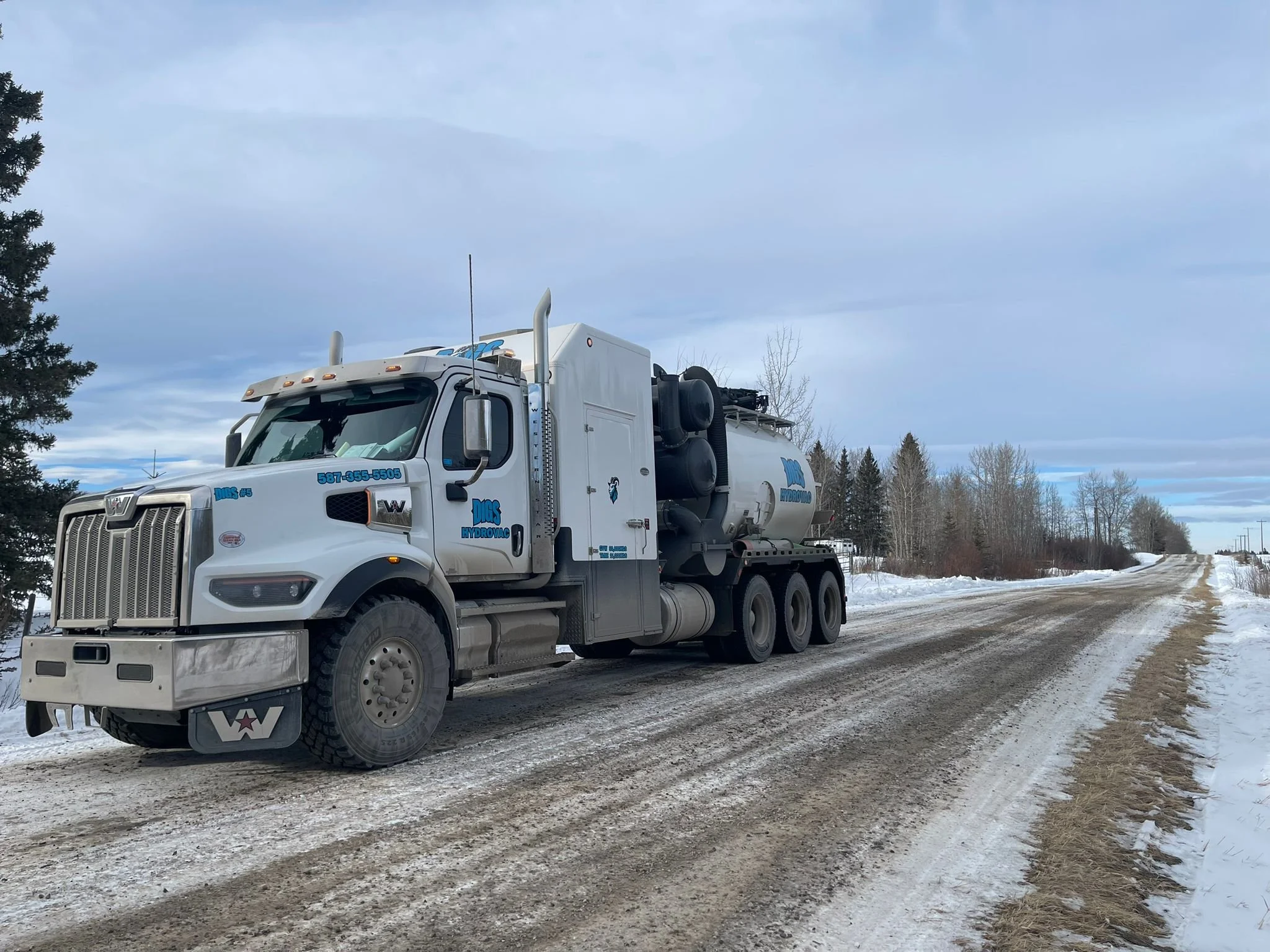 A large white fuel tanker truck parked on a snow-covered rural road with trees in the background and a cloudy sky.