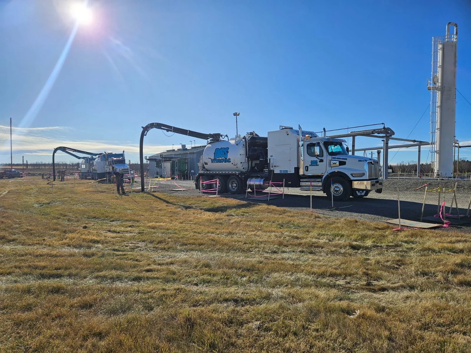 Two large vacuum trucks are parked outside a facility, with one spraying or extracting liquid into a tall storage tank on the right. The scene is set on a sunny day with a clear blue sky, and the area is marked off with pink and yellow caution tape.