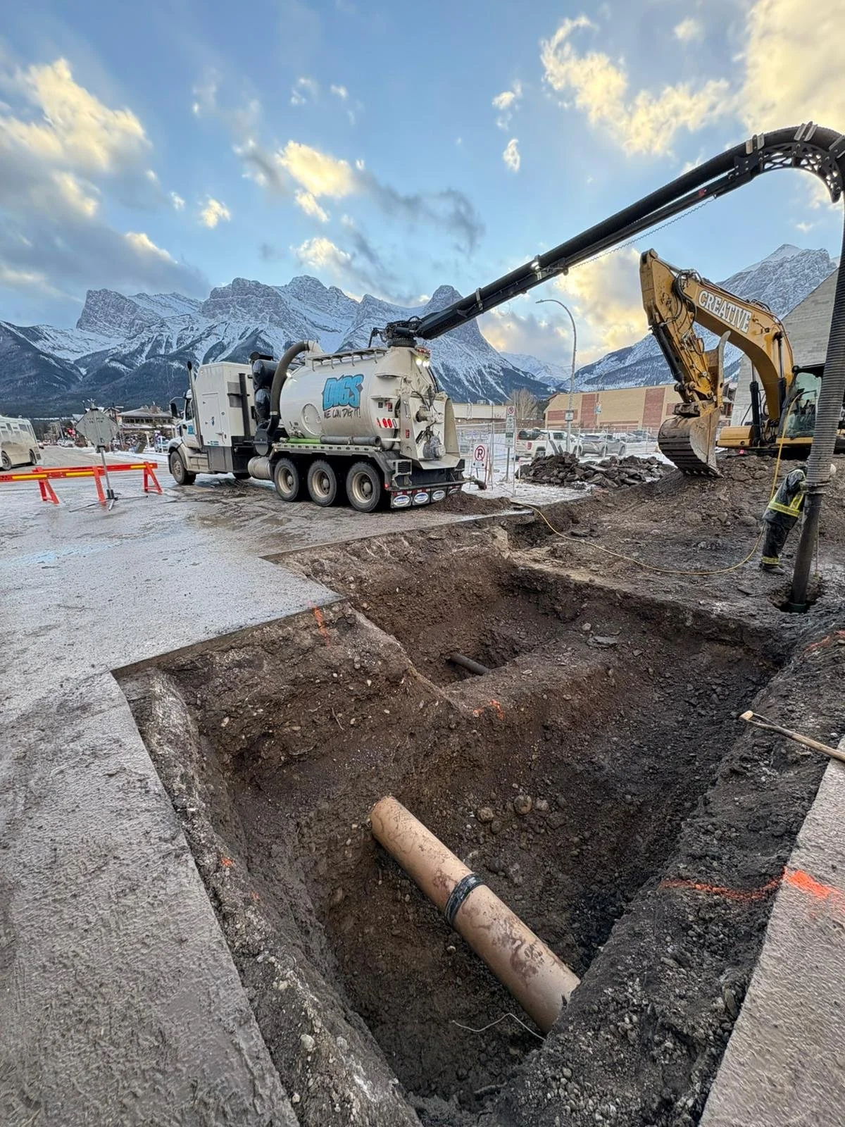 Construction workers installing underground piping in a trench with a vacuum truck and excavator, mountains in the background.