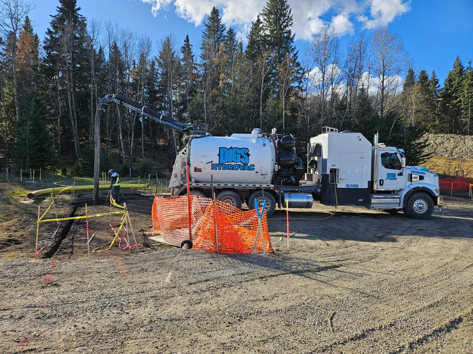 Construction site with a vacuum truck labeled 'DIGS HydroVac' parked on dirt, working near a tree where a worker in safety gear is using equipment. Trees and blue sky with clouds are in the background.