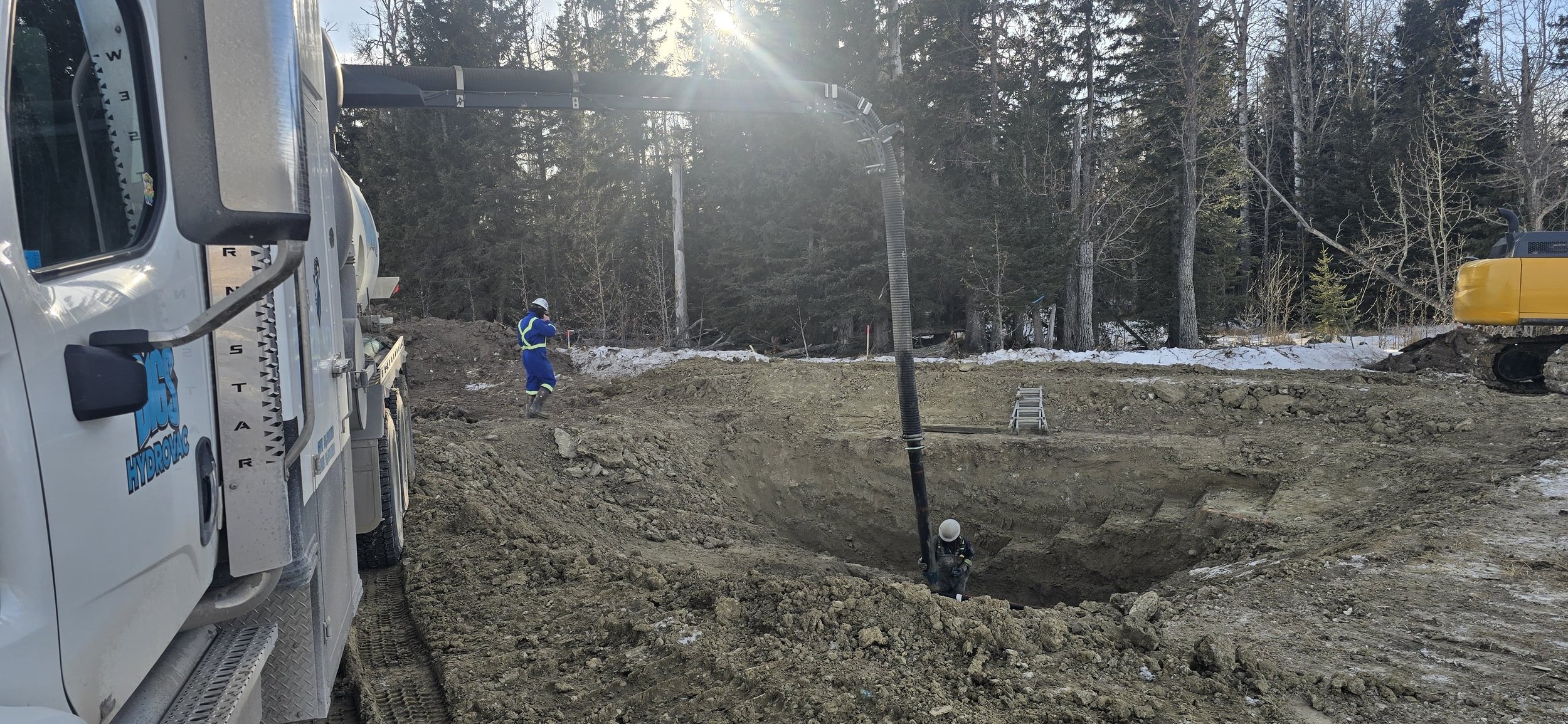 Construction site with two workers in safety gear and a hydraulic excavator in the background, surrounded by a wooded area, snow on the ground, and a large dirt excavation.