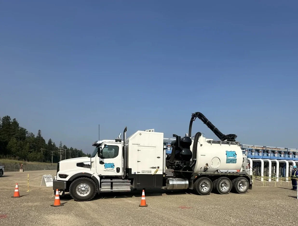 A vacuum truck with a removable tank and hose, parked on a gravel lot with orange cones and a construction worker nearby. The truck has logos and is set against a clear blue sky with trees and industrial structures in the background.
