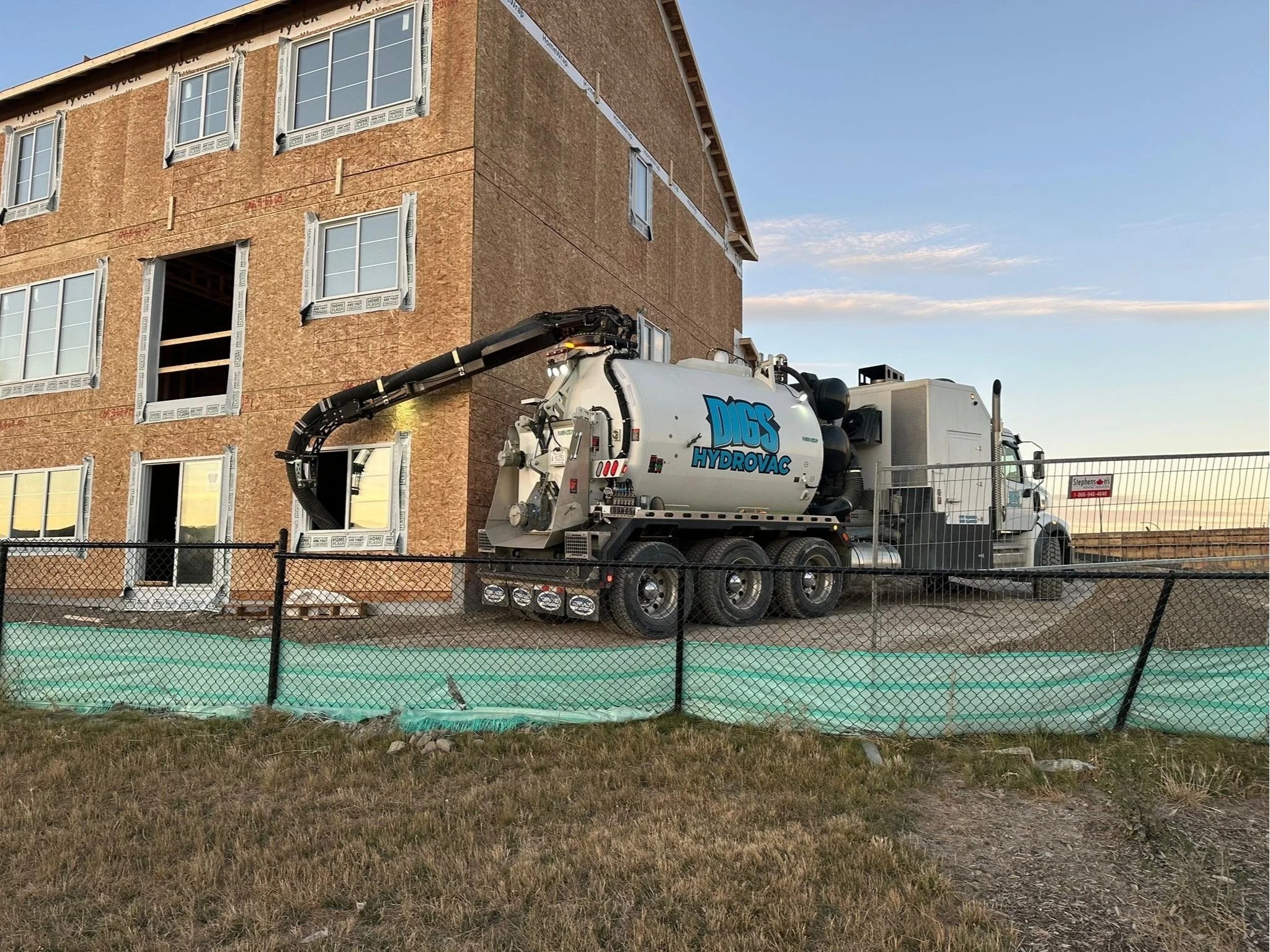 A construction site with a partially built multi-story wooden building and a large vacuum truck labeled 'Digs Hydrovac' parked on a level concrete surface, surrounded by a chain-link fence.