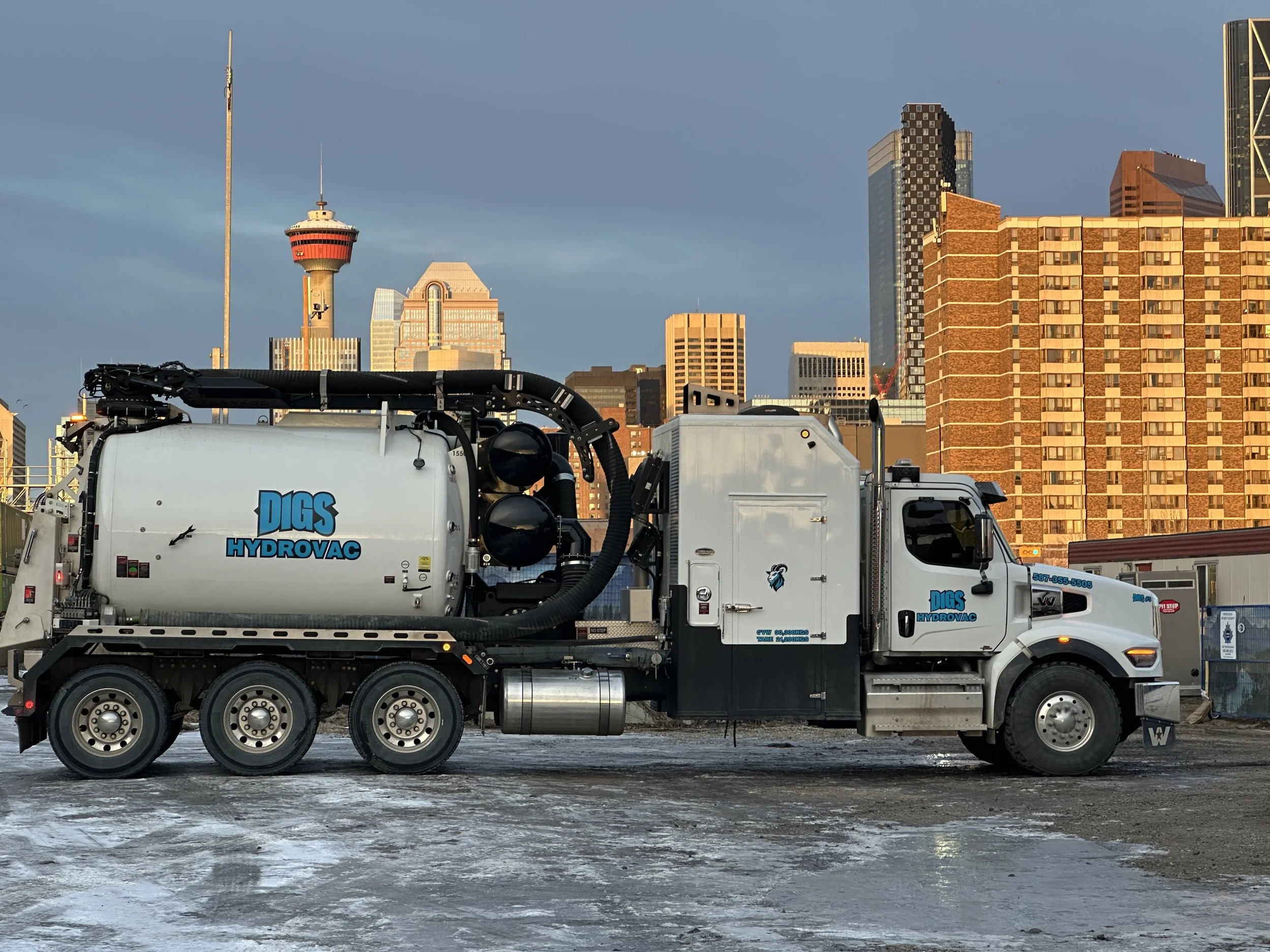 A white hydro excavation truck labeled 'DIGS HYDROVAC' parked on a snowy surface in an urban area with a city skyline, including the Calgary Tower, in the background.