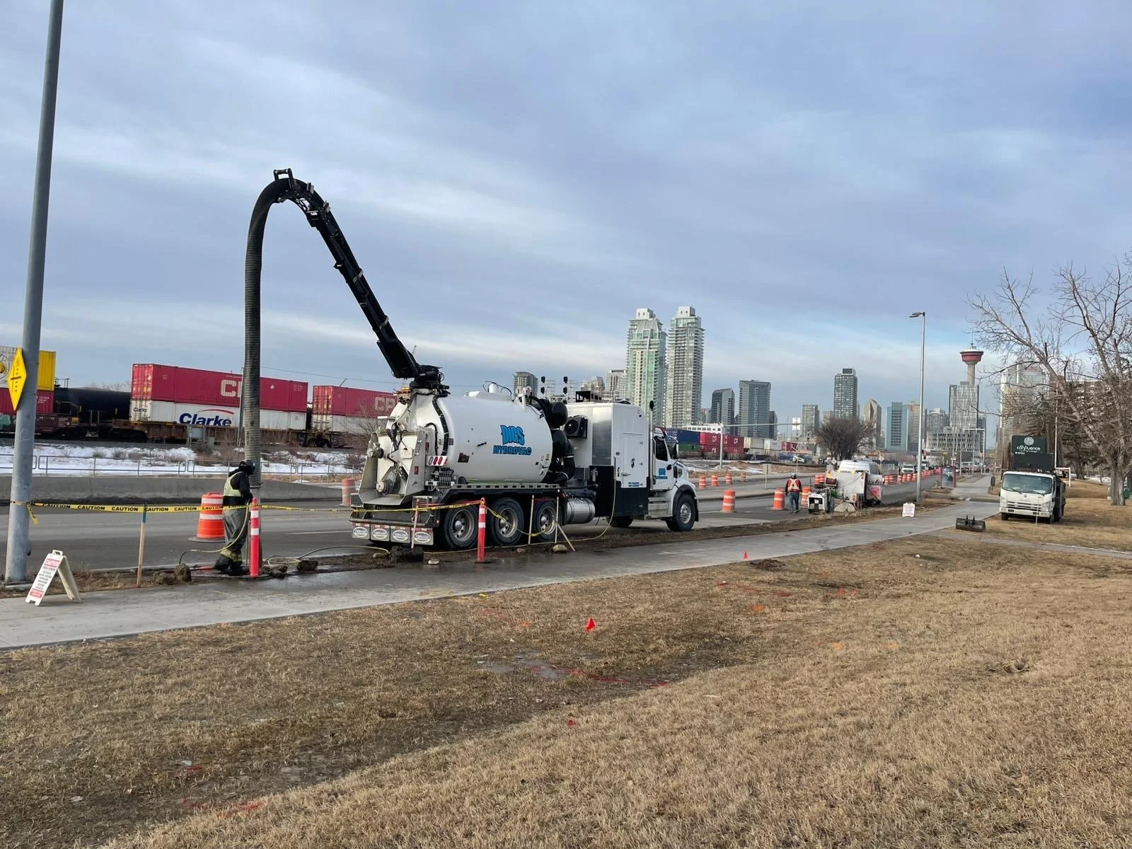 Construction workers and a vacuum truck working on a sidewalk, with the city skyline in the background, in an urban area with some trees and traffic cones.
