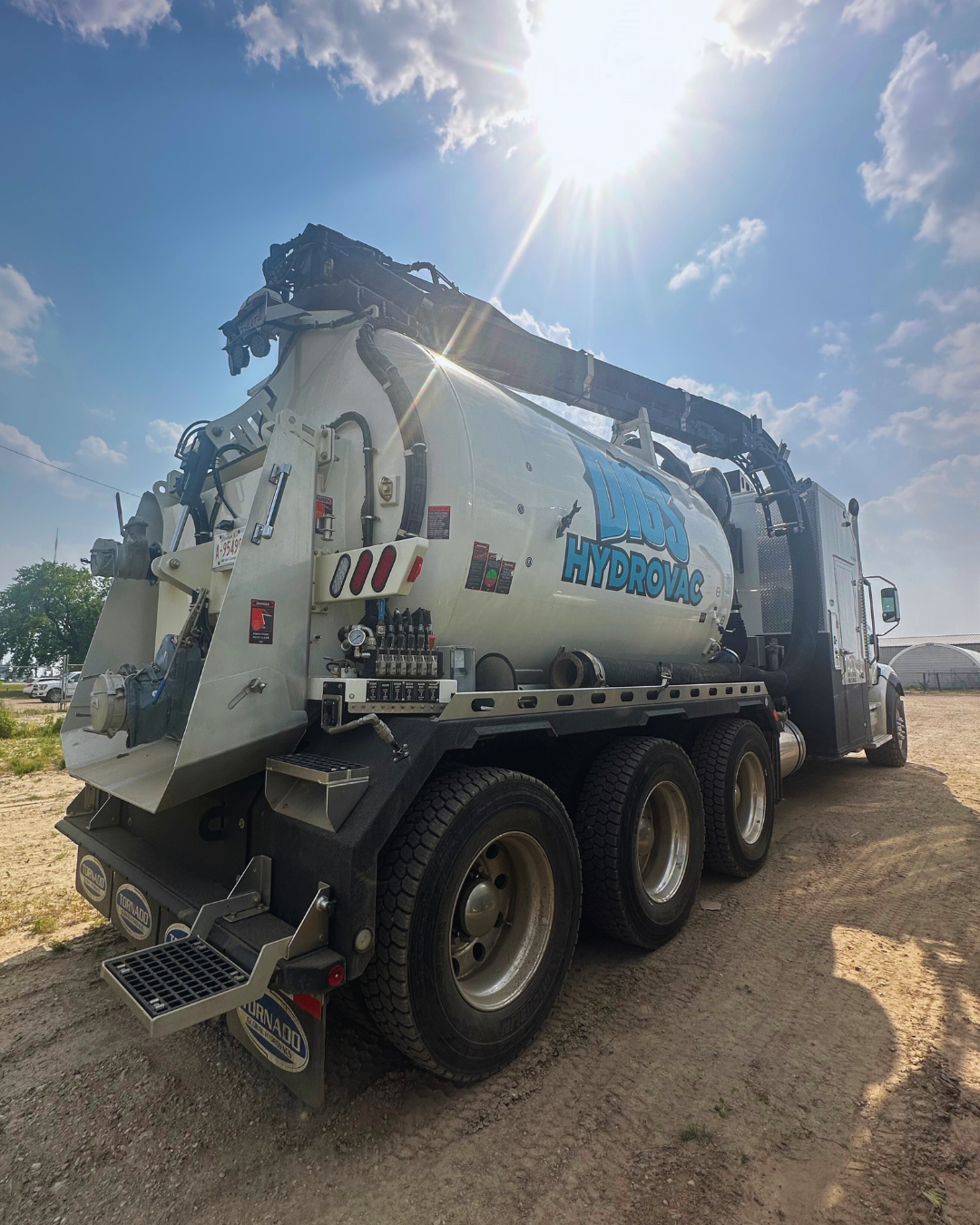 A large Hydrovac truck with a white tank and black crane arm parked on a dirt lot under a bright sun and partly cloudy sky.