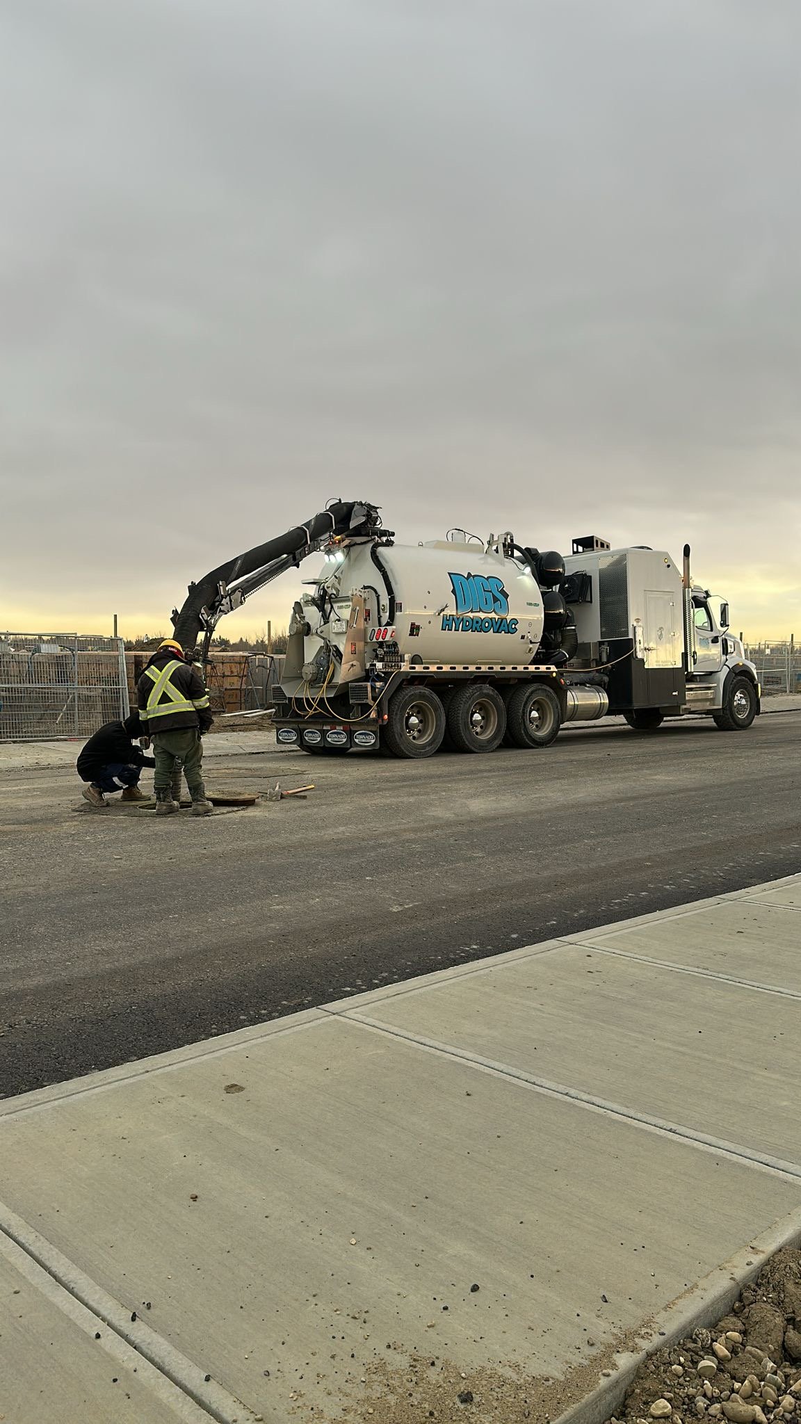 Construction workers in safety vests working near a large HydroVac vacuum truck on a road construction site.