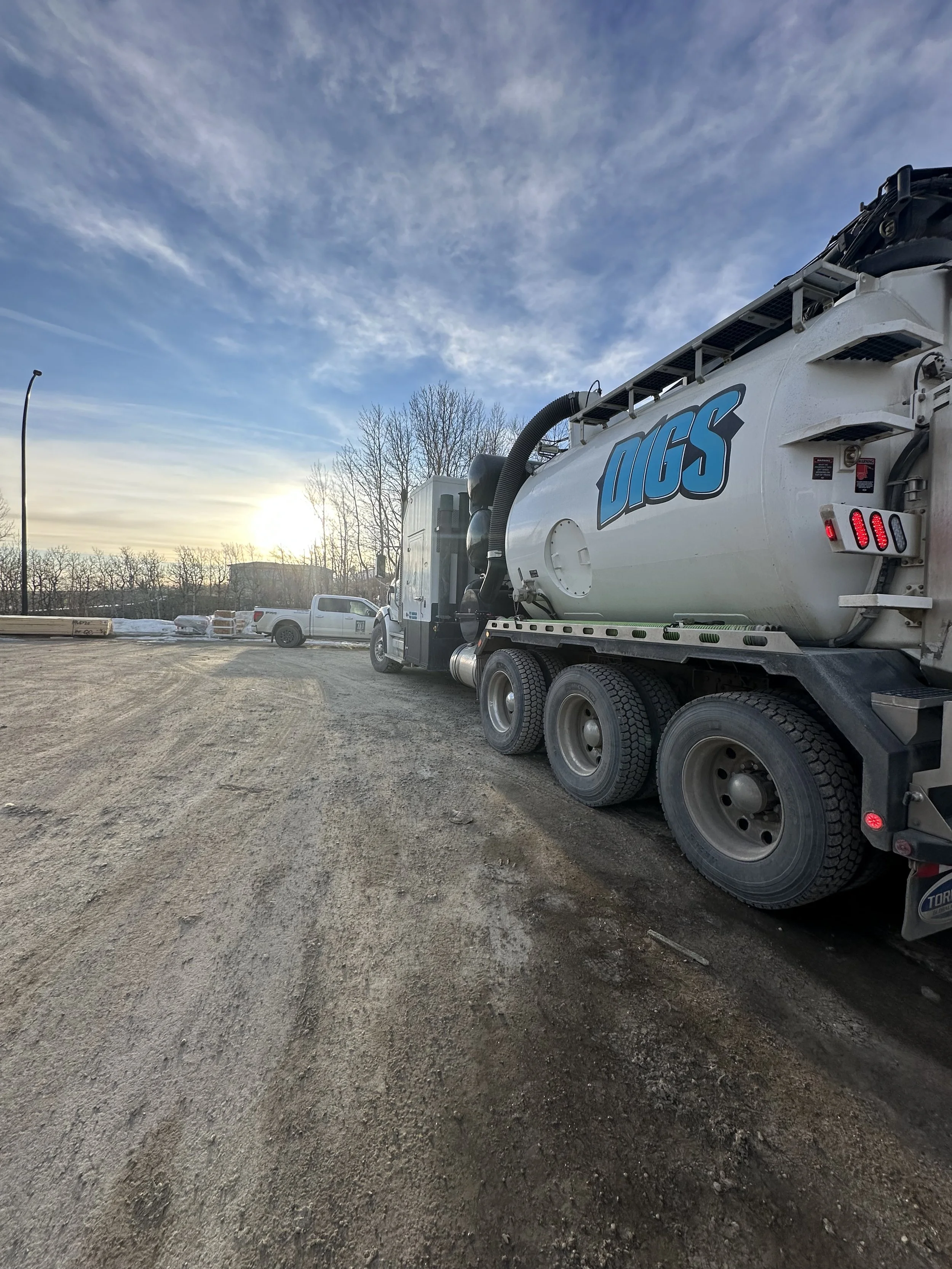 A large white vacuum truck with the words 'DIGS' on the side, parked on a gravel lot during sunrise or sunset, with bare trees and several other vehicles in the background.