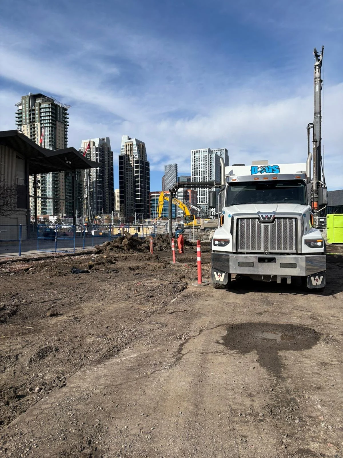 Construction site with a large dump truck, dirt piles, and modern high-rise buildings in the background under a partly cloudy sky.