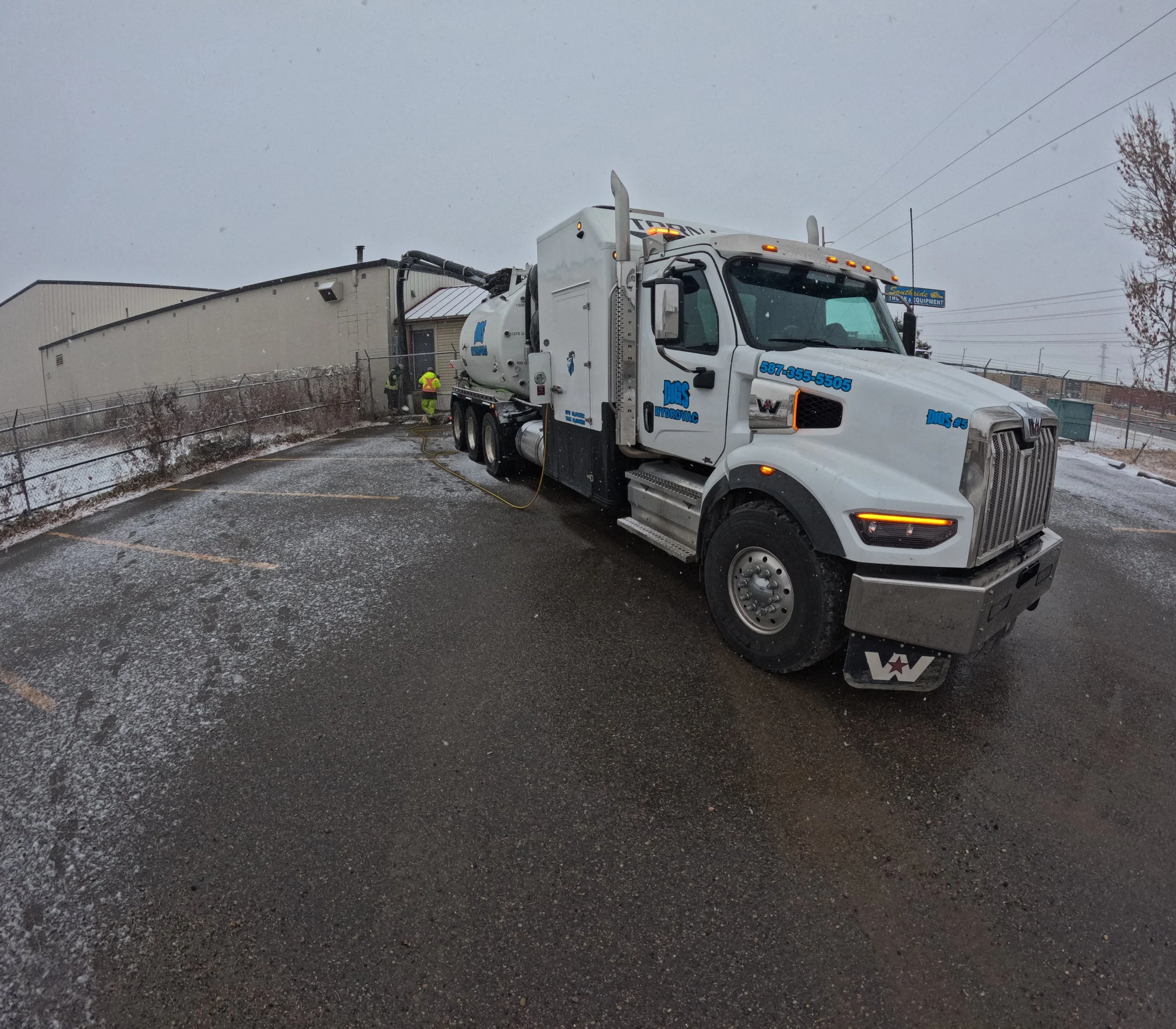 A concrete mixer truck parked outside a warehouse building on a snowy and icy day, with workers in safety vests near the warehouse entrance.
