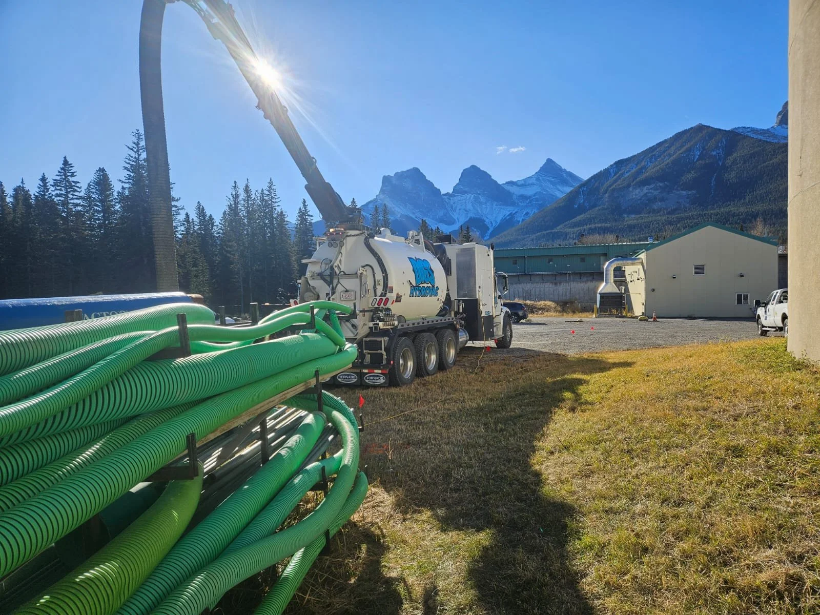 Construction site with large green hoses, a water truck with blue logo, a crane, a building, and mountain scenery in the background.