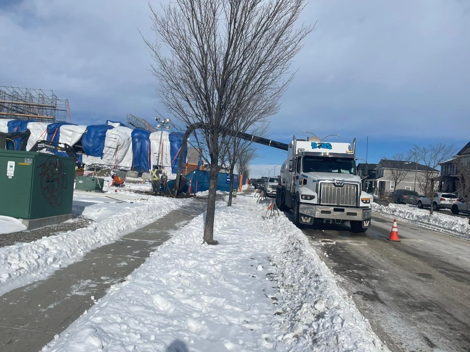 A snow-covered sidewalk and street with a tree in the foreground, a construction crane and workers near a building covered with tarps, and a utility truck with a cherry picker on the street, with houses and parked cars in the background.