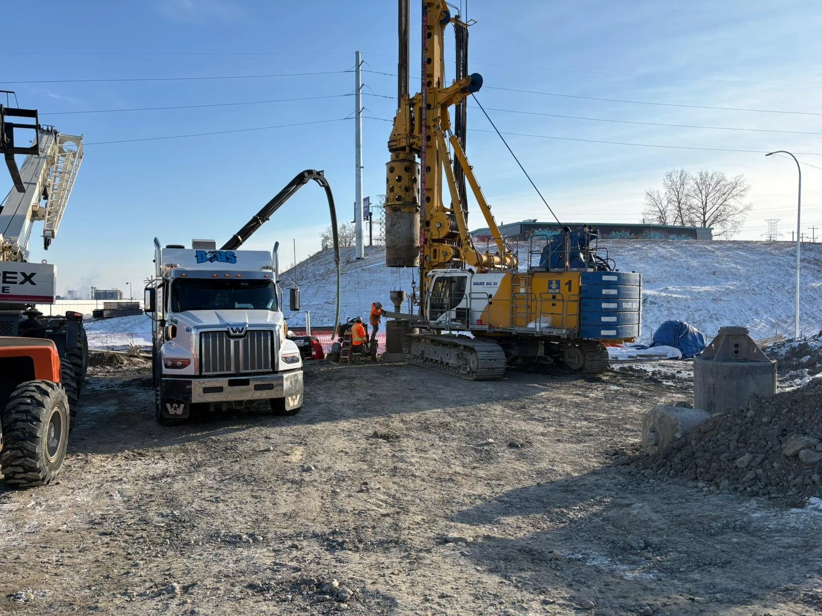 Construction site with a large yellow drilling machine, a white truck with a black boom, and workers in orange safety vests. Snow is on the ground and background hills, with trees and utility poles.