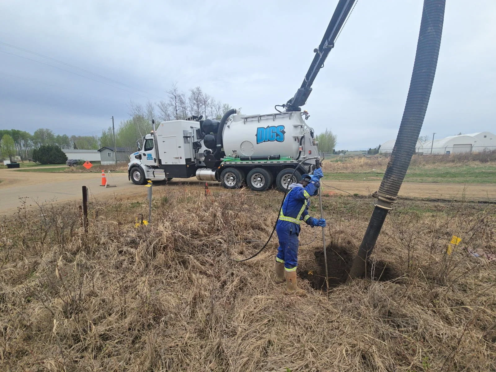A worker in a blue uniform and safety gear operating a large industrial vacuum truck labeled "DIGS" to extract liquid from a drilled hole in the ground outside, with a cloudy sky overhead.