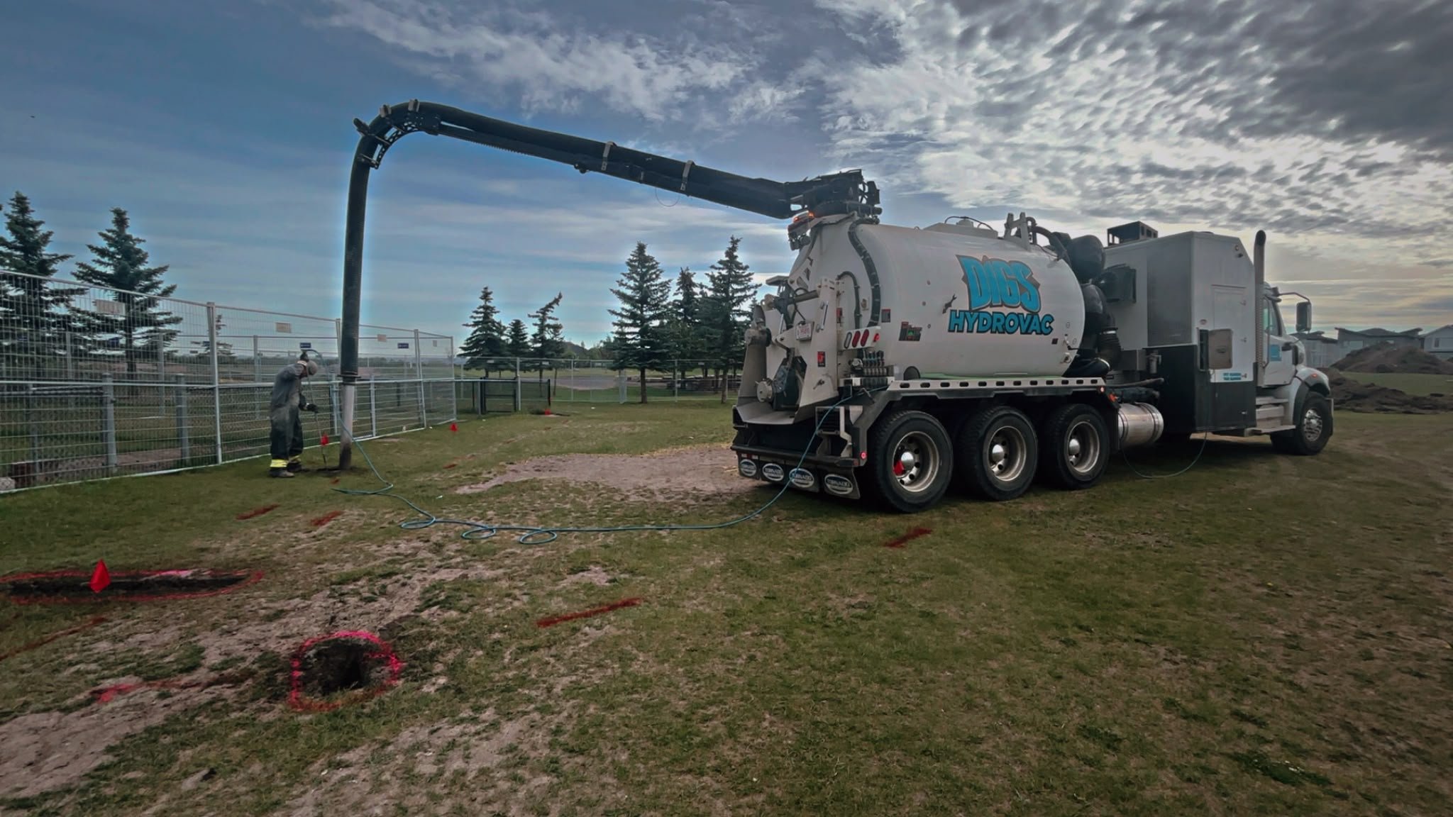 DIGS Hydrovac tanker truck with a long arm pouring water into a confined hole during hydro excavation, with two workers nearby, fencing, and trees in the background.