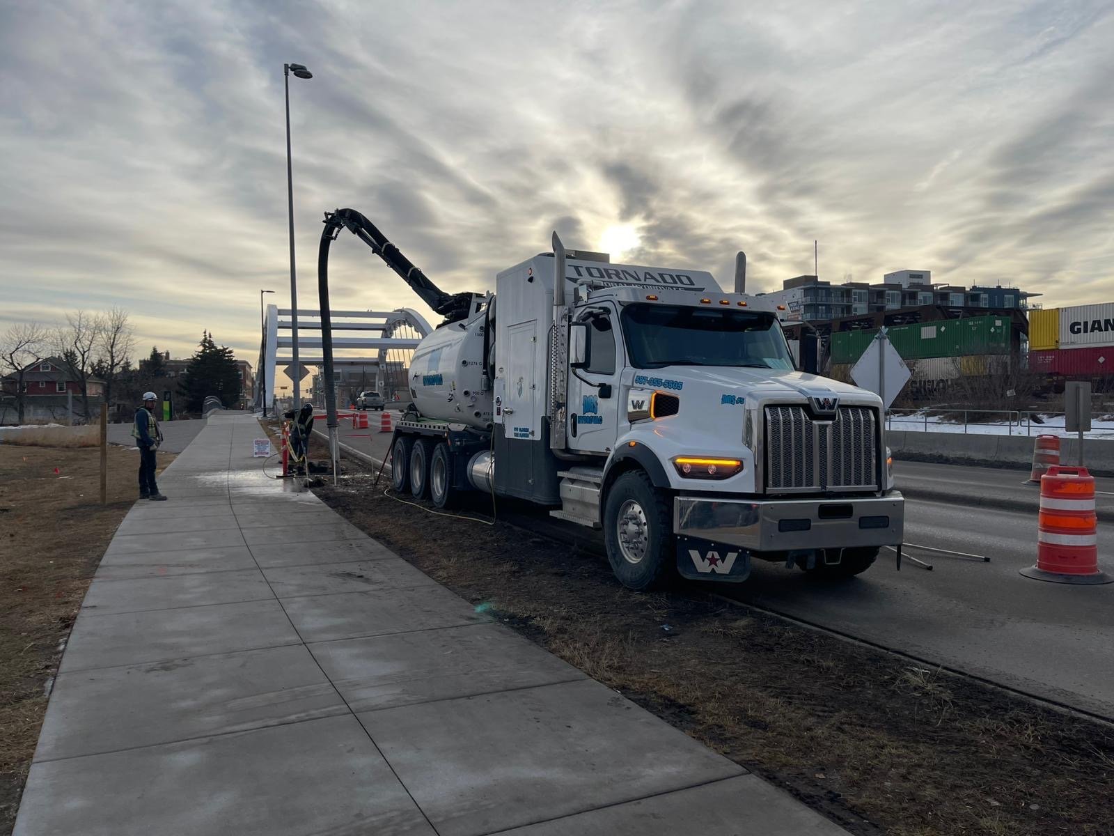 A large white vacuum truck parked on a sidewalk construction site, with construction workers and orange safety cones nearby, under a cloudy sky.