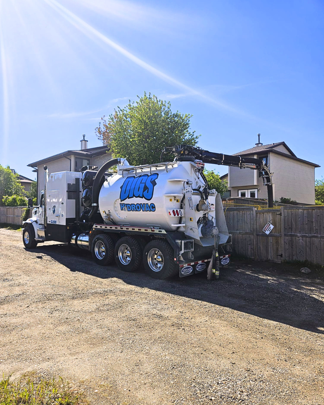 DIGS Hydrovac vacuum truck parked on a gravel lot, with a house and fence in the background, under a clear blue sky with bright sunlight.