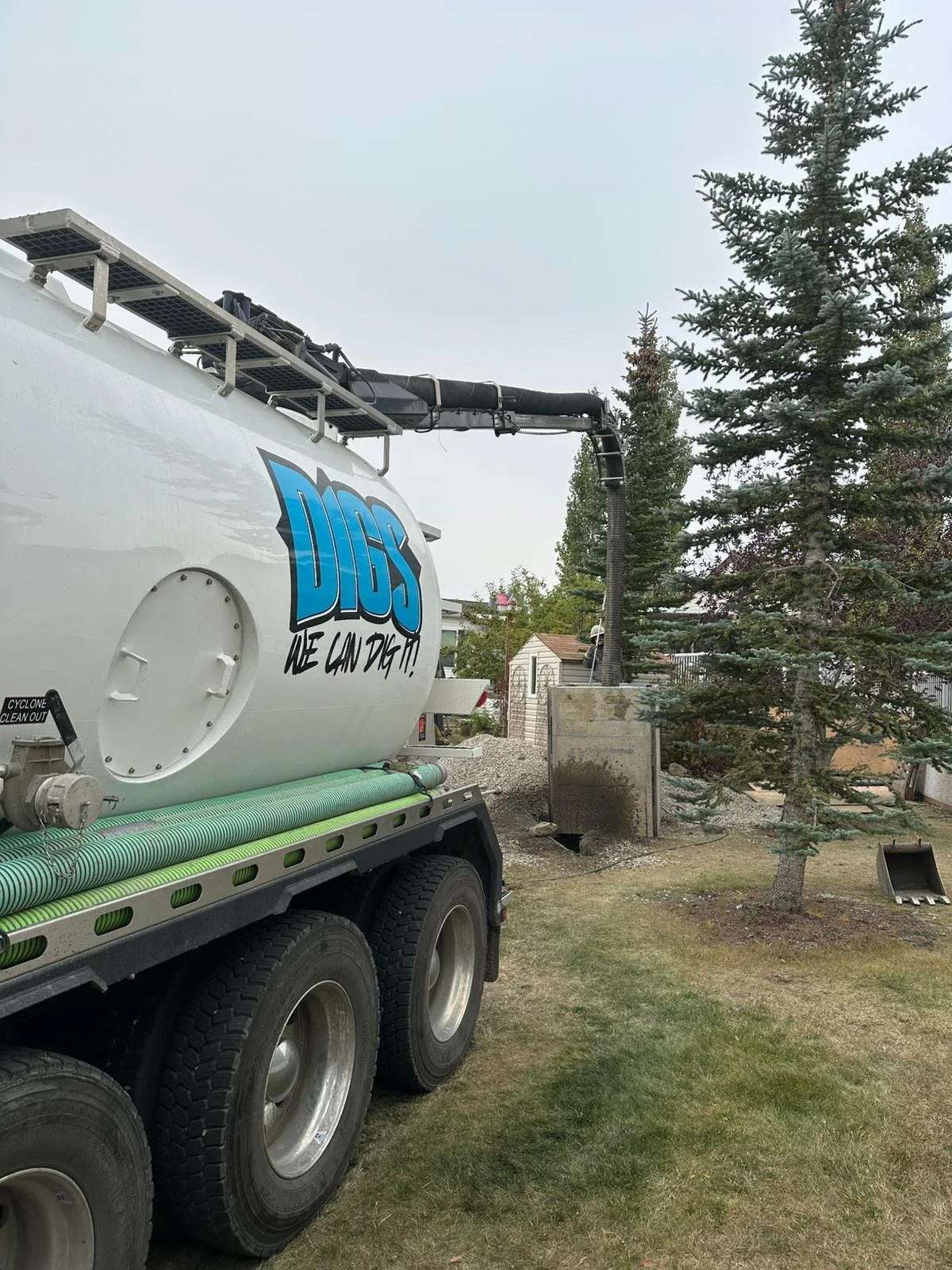 A large mobile machine with a tank labeled 'DIGS We Can Dig It!' is positioned on a grassy yard, connected to a black pipe that leads to an underground excavation site near a tree, with a small structure in the background.