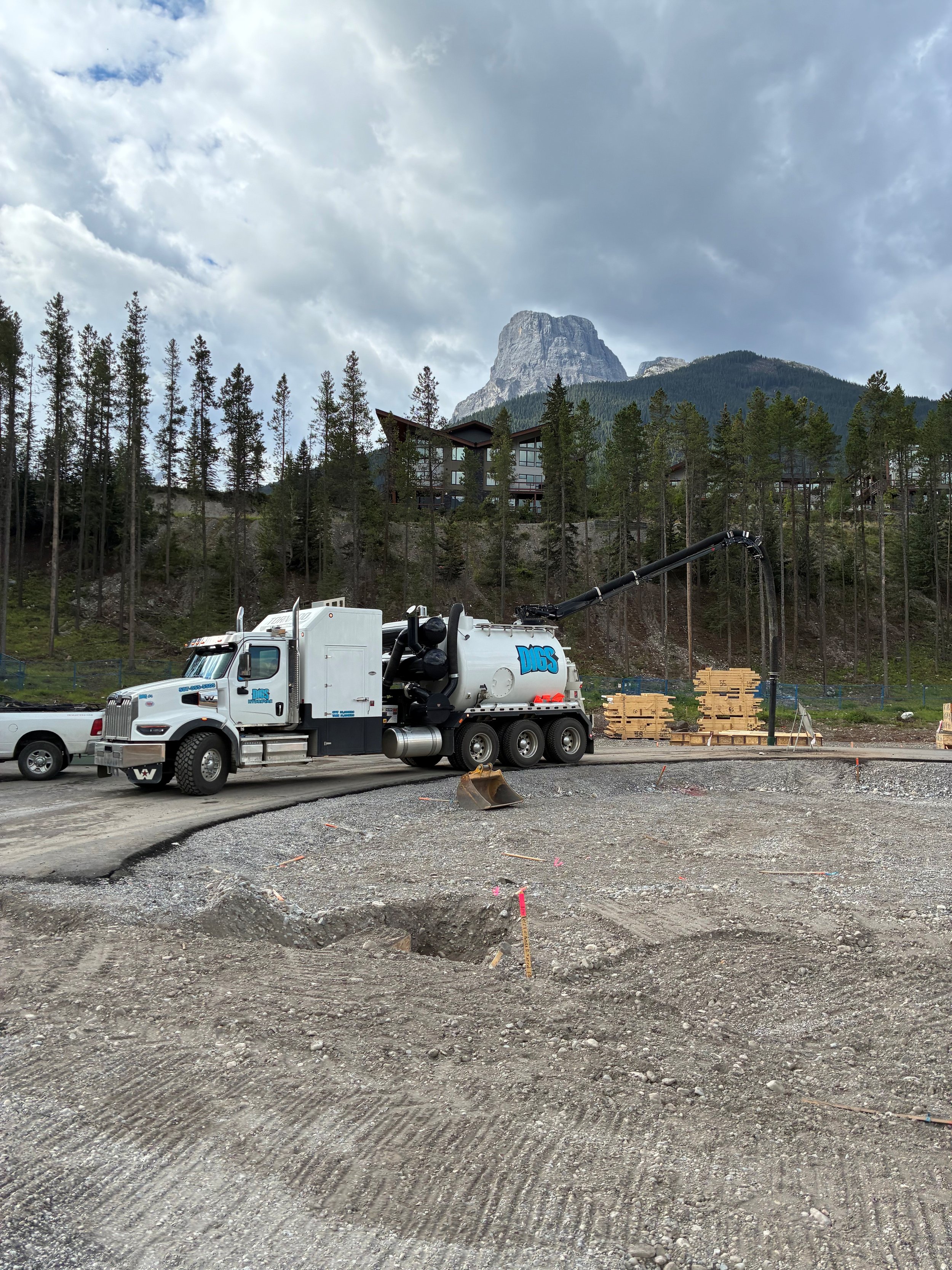 A construction site with a large white dump truck and a truck-mounted vacuum or pump, with a mountain and trees in the background.