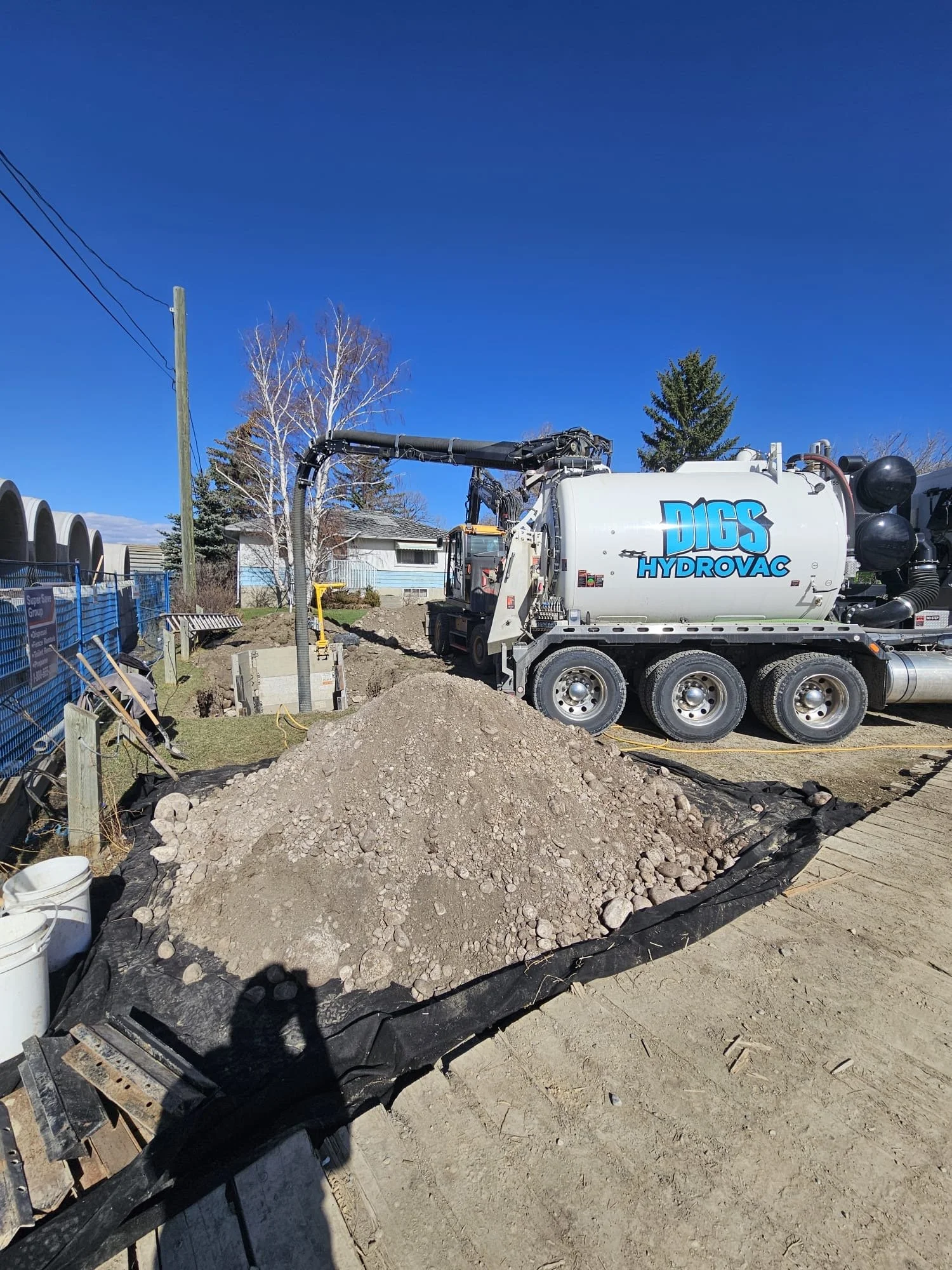 Construction site with a large white vacuum truck labeled DICS Hydrovac, dirt piles, a black plastic sheet on the ground, and construction materials. There are trees, a small house, and a blue sky in the background.