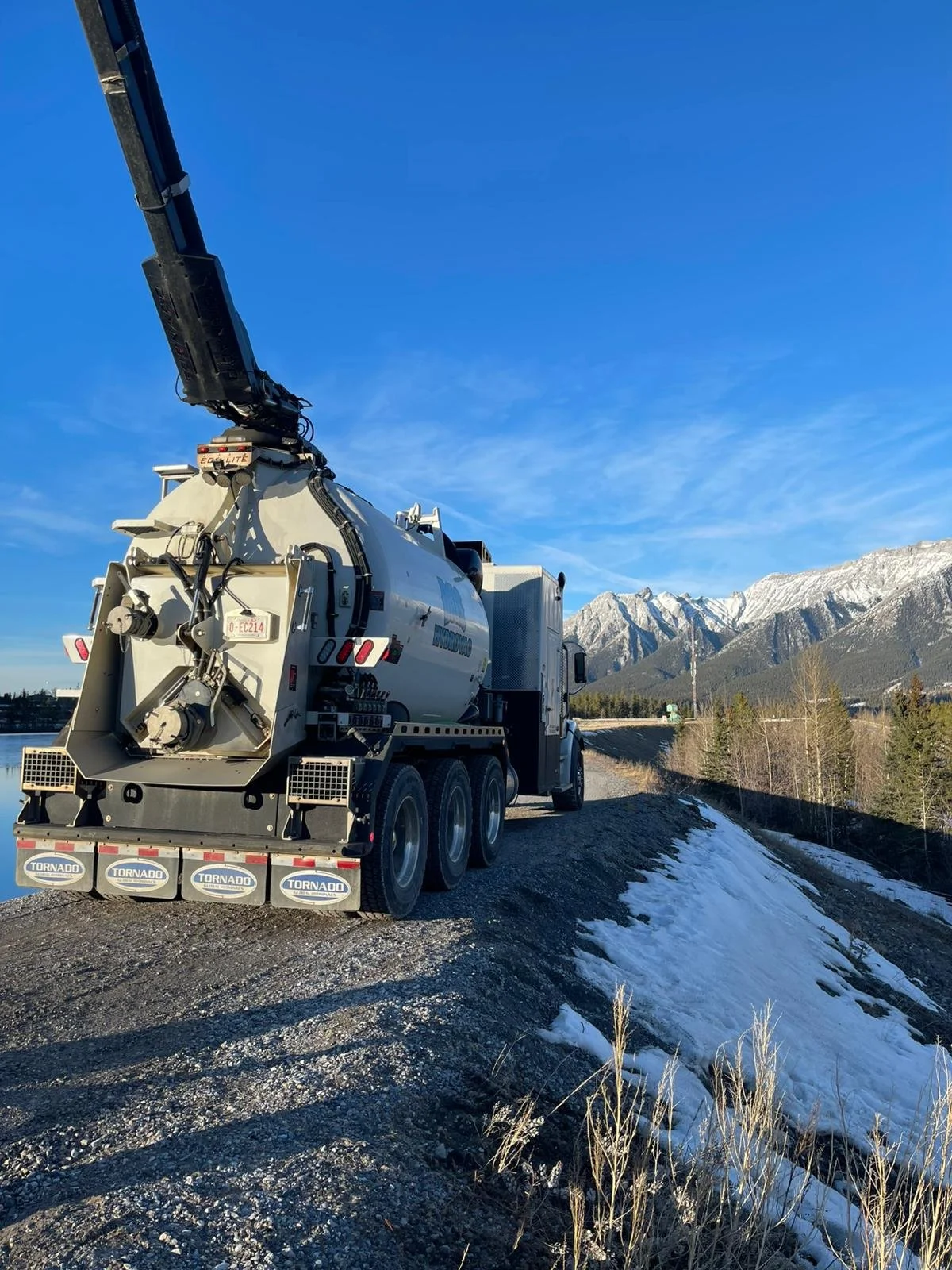 A vacuum truck with a large cylindrical tank parked on a gravel road with snow, mountains, trees, and a clear blue sky in the background.
