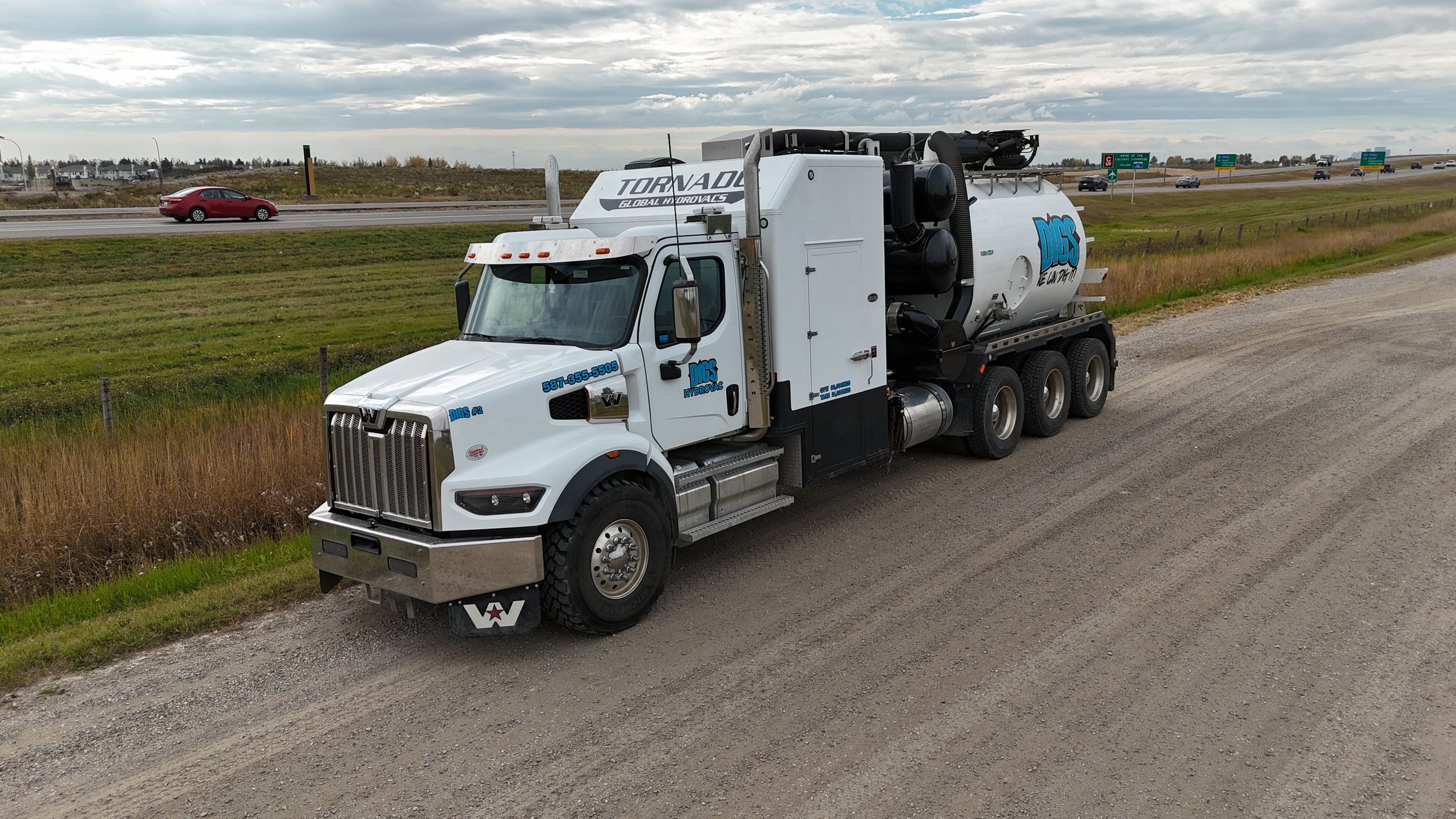 A Hydrovac truck for DGS Hydrovac parked on a roadside with a grassy field and highway in the background on a cloudy day.