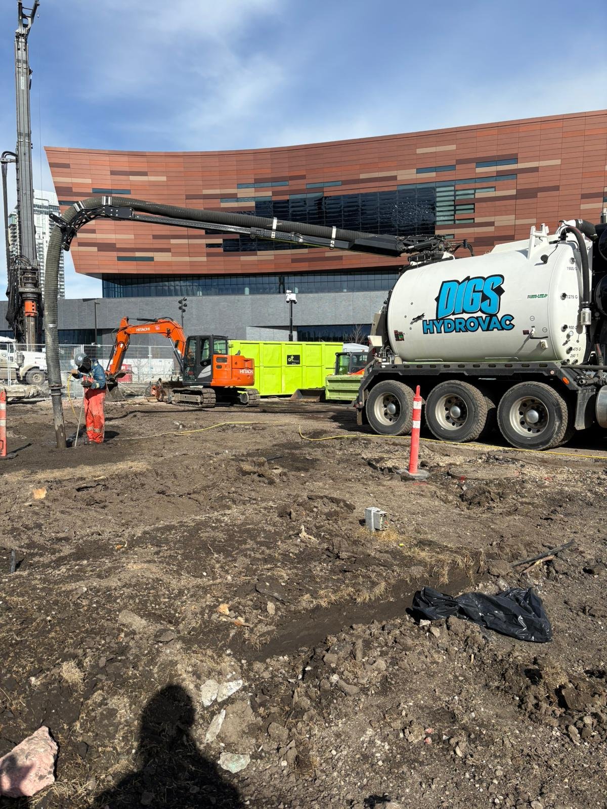 Construction site with dirt ground, a worker, heavy machinery including a vacuum truck labeled 'Digs HydroVac', a small orange excavator, and a yellow container in front of a modern building with brown and black panels.