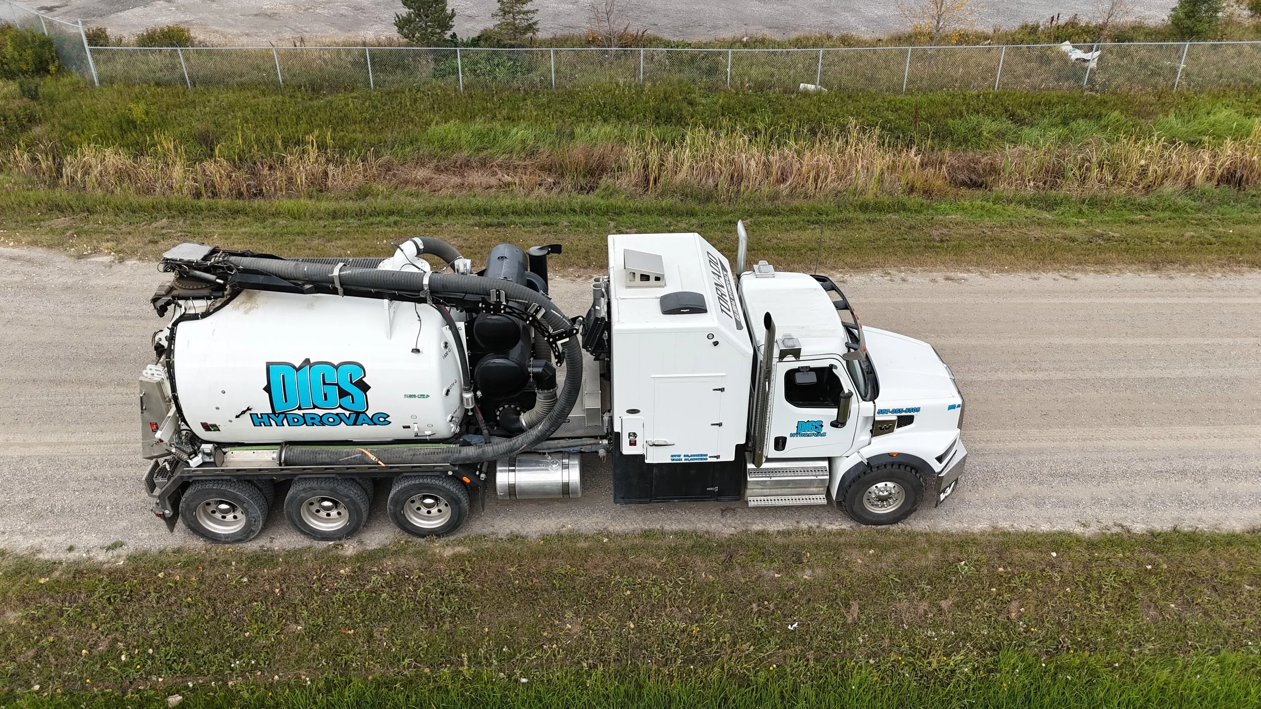A large white vacuum truck with the logo "DIGS HYDROVAC" is parked on a gravel road, with grassy fields and a chain-link fence in the background.