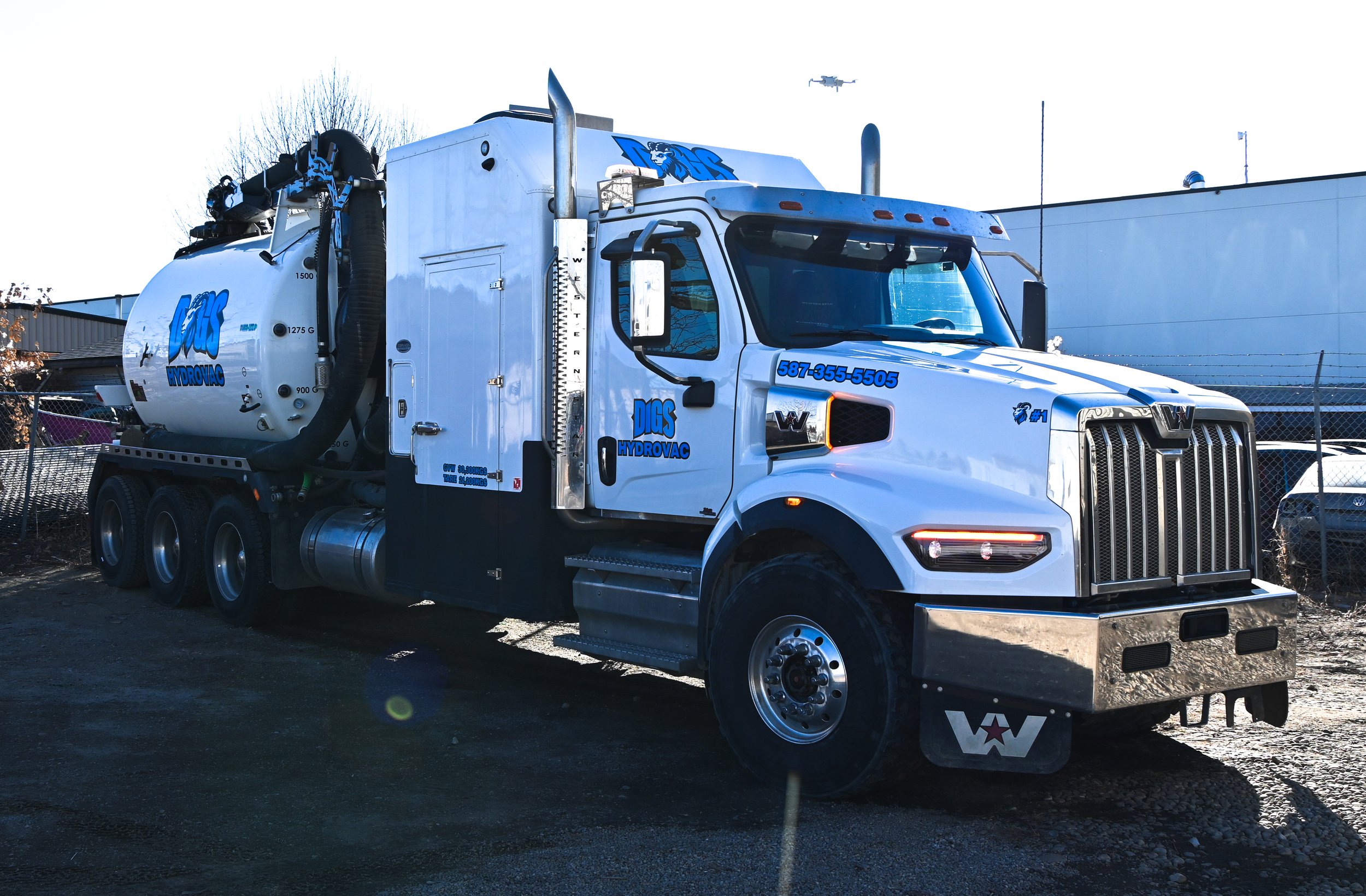 A white water delivery truck with blue markings and a large tank on the back, parked outdoors near a chain-link fence and industrial buildings.