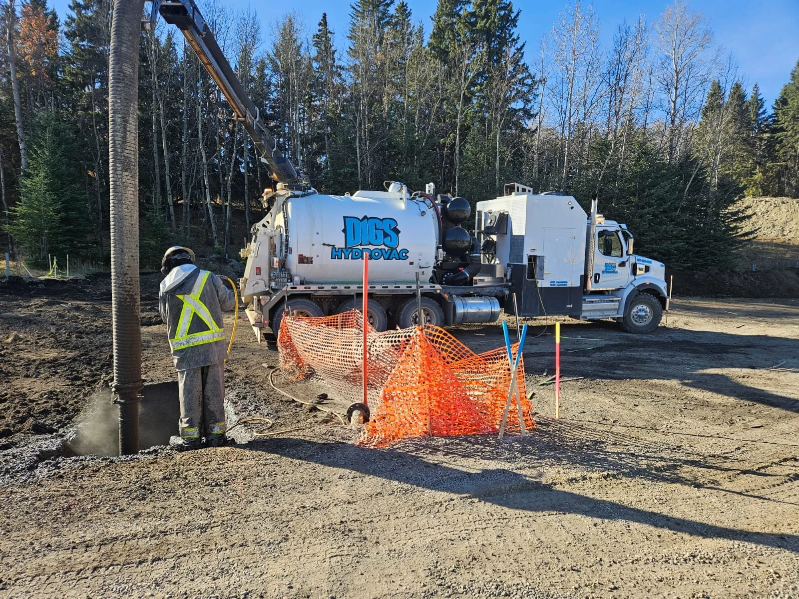 Worker in safety vest and helmet near a construction site with an orange safety fence, a large white hydro excavator truck with the label 'Digs HydroVac,' and a background of trees.