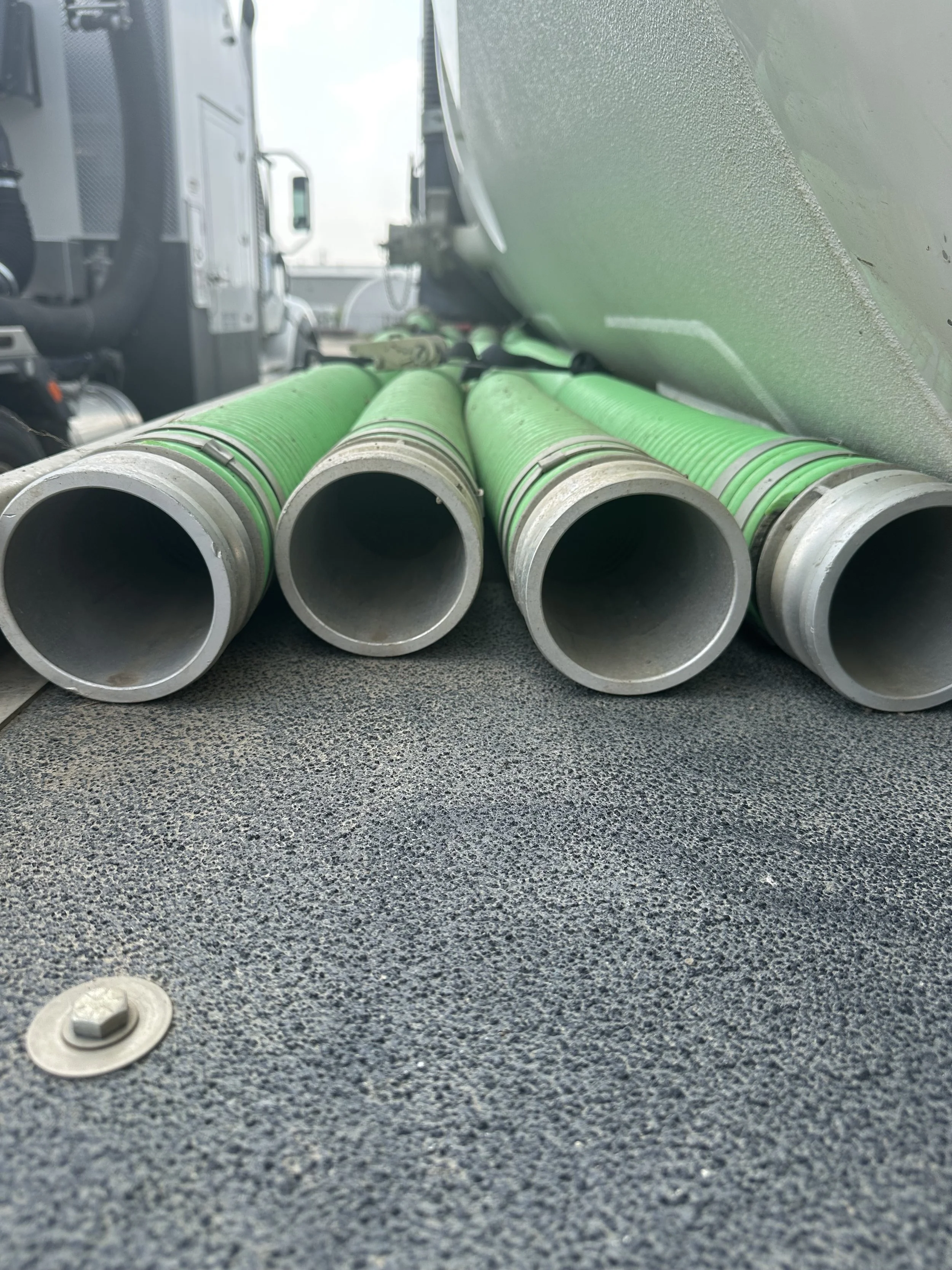 Four green pipes with metal ends are lying parallel on a textured black surface near a large truck or machinery.