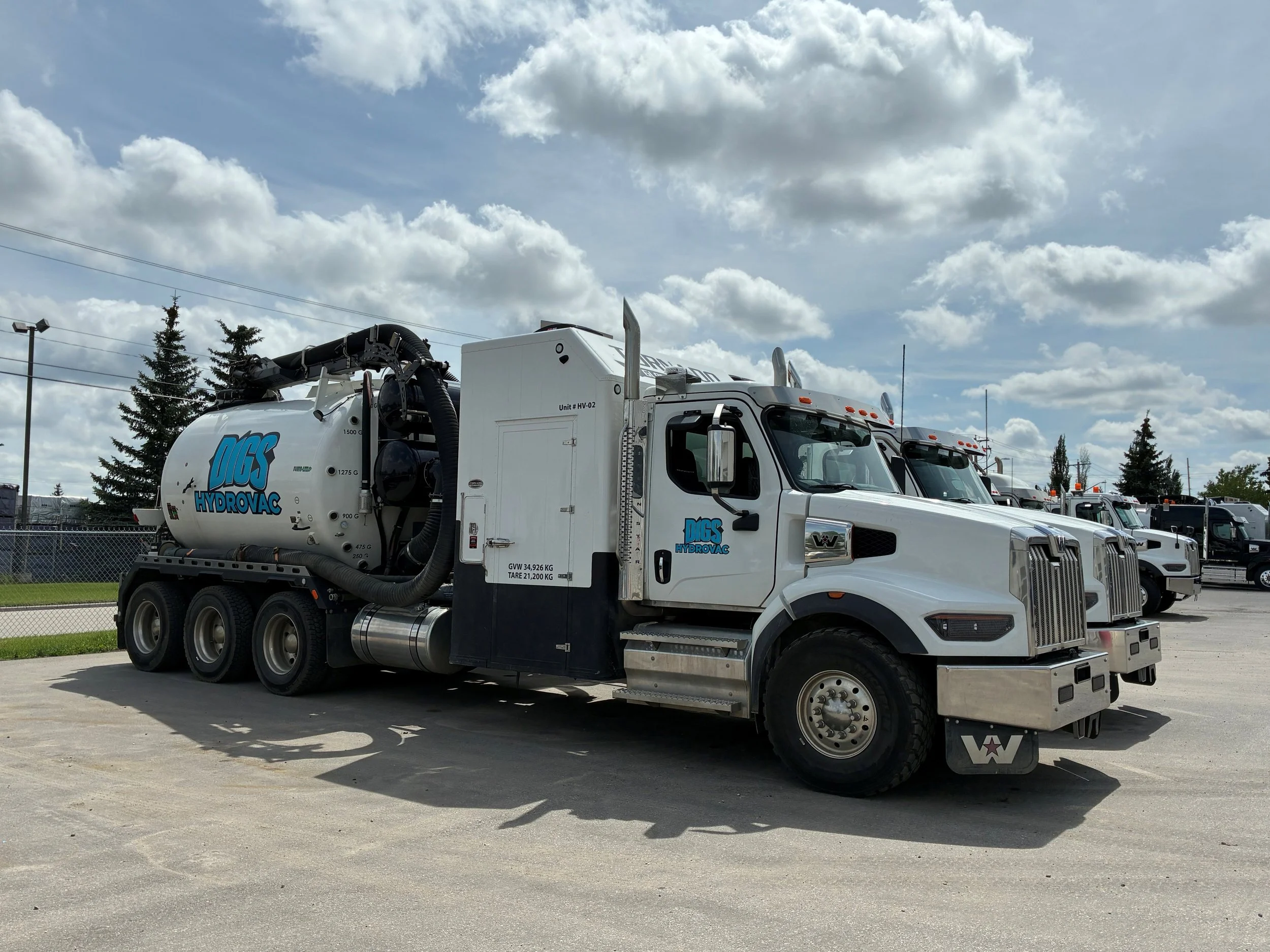A large white industrial vacuum truck with the logo 'Digs HydroVac' parked in a lot with other trucks under a partly cloudy sky.