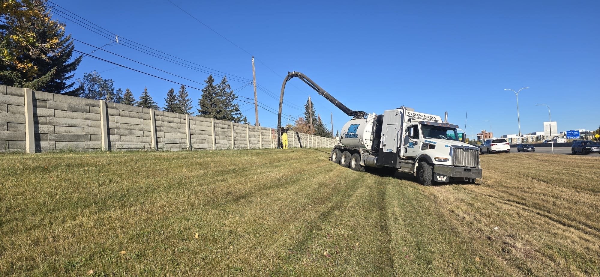 A white vacuum truck with the word 'TORNADO' on it and a large hose attached, vacuuming or cleaning an area near a tall concrete fence, with a worker in yellow in the background and a busy highway with cars and buildings in the distance under a clear blue sky.