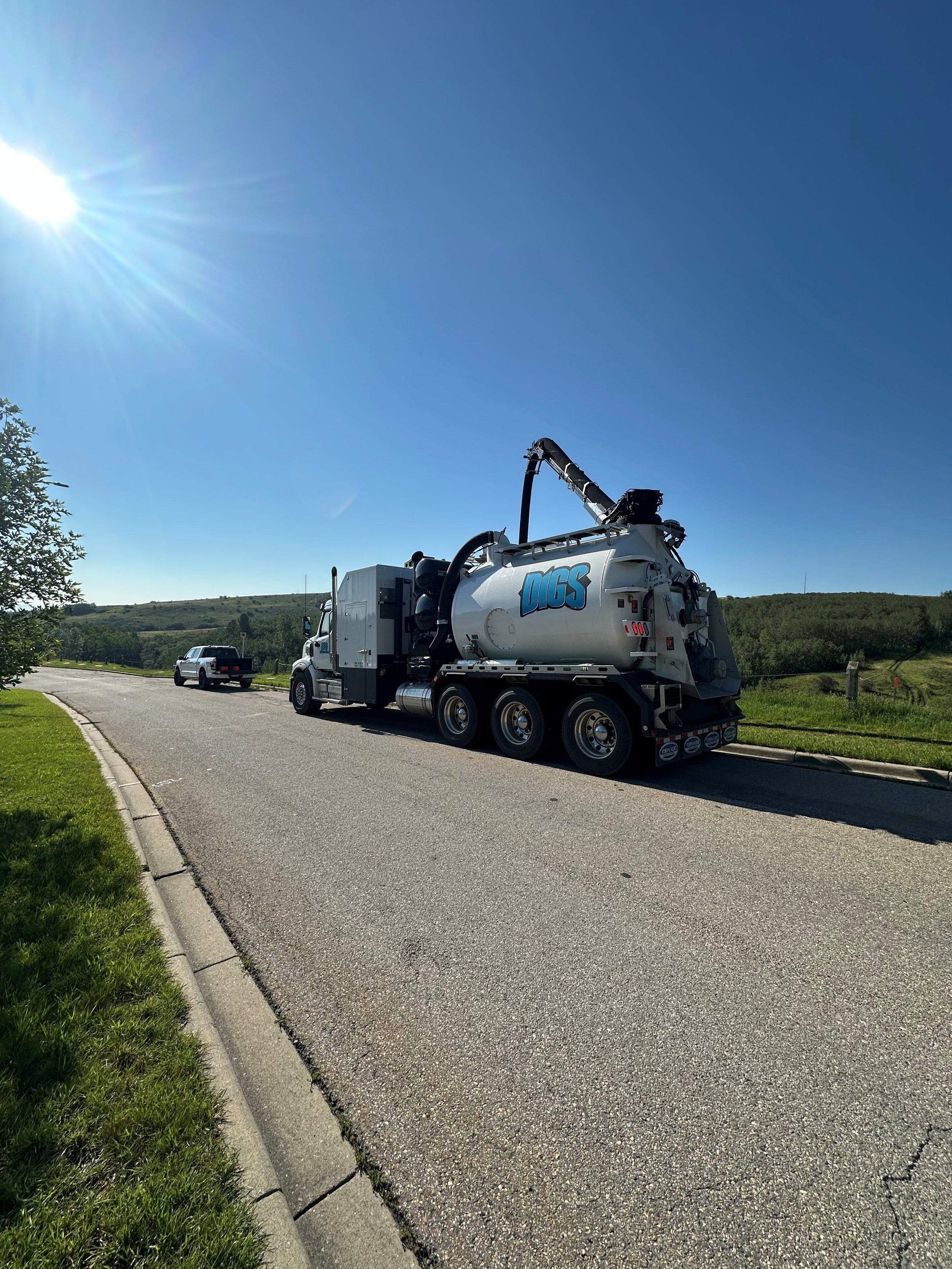 A large vacuum truck with the word 'DOGS' on the side is parked on a rural street under a bright, sunny sky, with a white SUV vehicle further down the road and greenery surrounding the area.