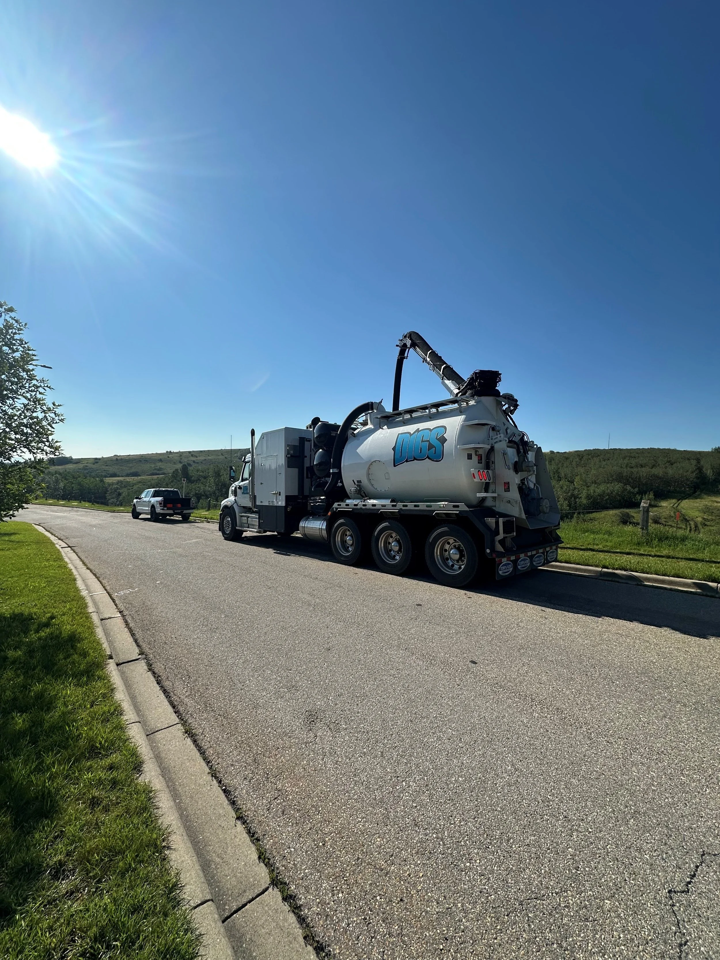 A vacuum truck with the word "DUGS" on the side parked on a rural road with grassy fields and hills in the background under a bright blue sky with the sun shining.