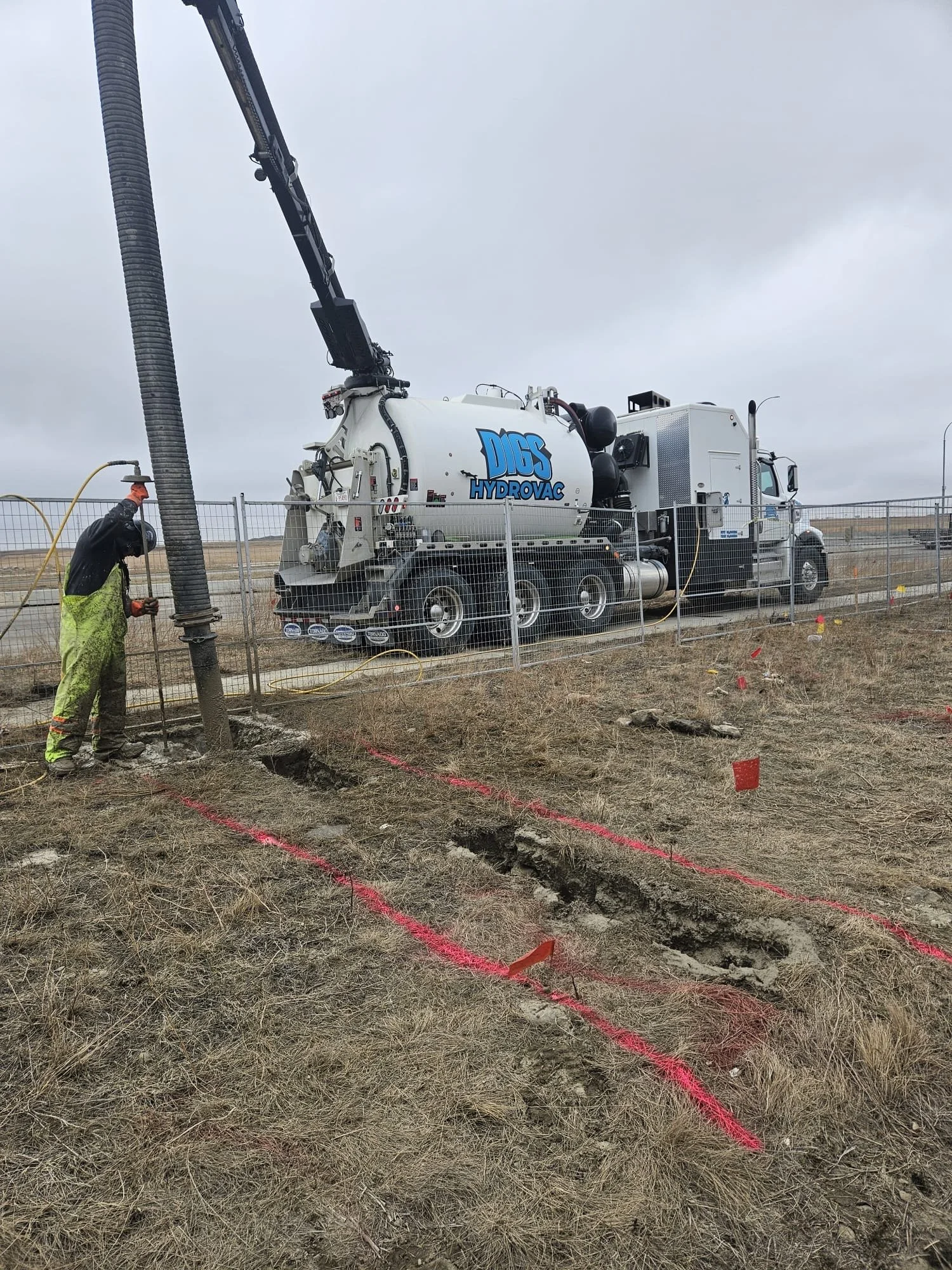 Worker operating a vacuum excavator truck labeled DICS Hydrovac, digging narrow trenches in dry grass field with marked boundaries.