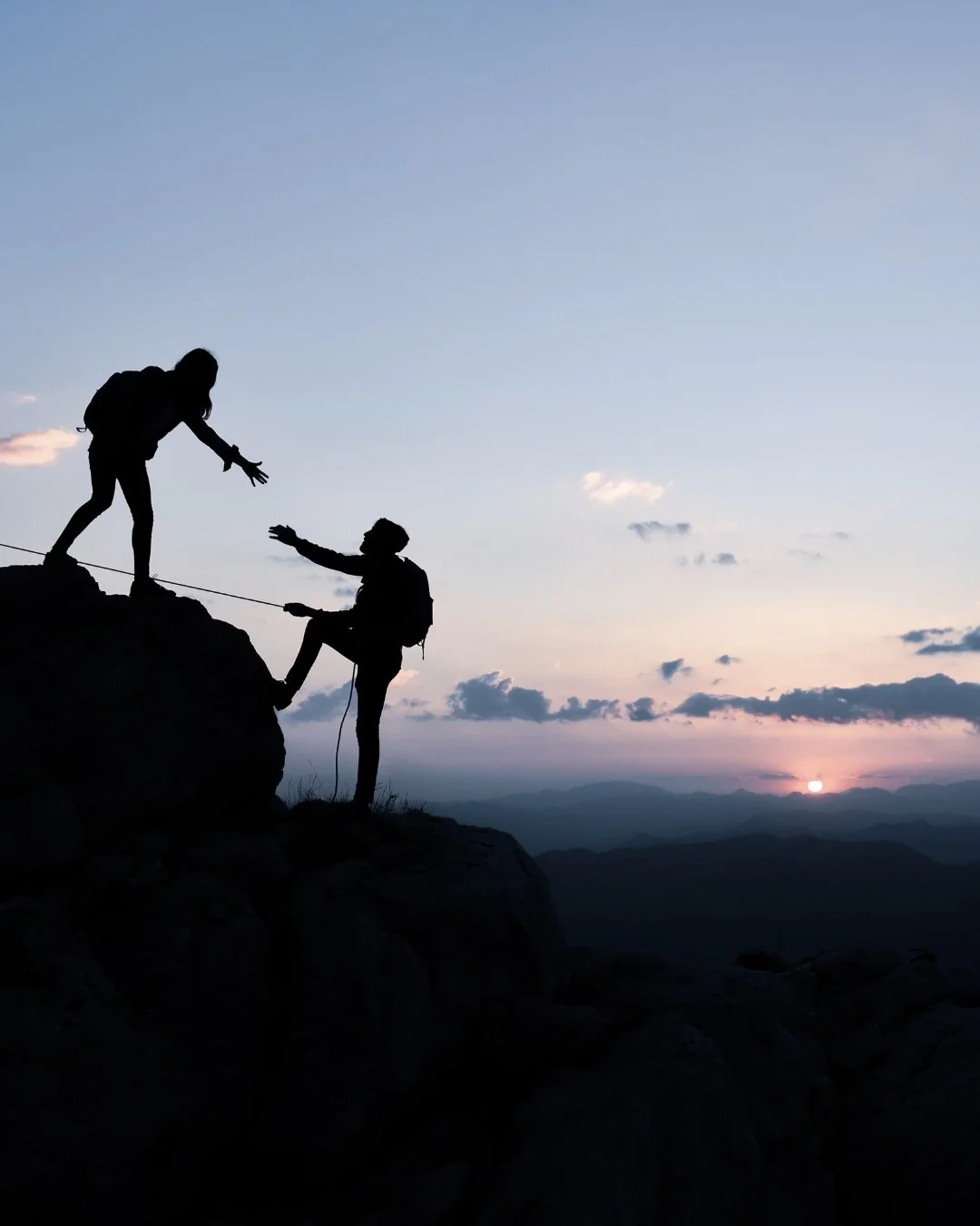Silhouette of one hiker helping another climb a mountain at sunrise, symbolizing how executive coaching provides guidance, support, and leadership growth