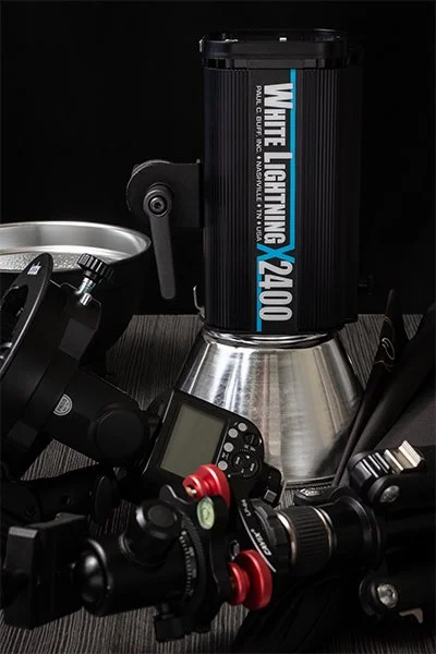 A collection of camera equipment, including a studio strobe, a camera, a lens, and a monitor, arranged on a dark surface.