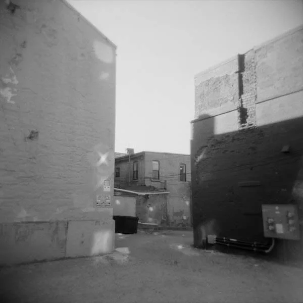 Black and white photo of an alleyway between brick buildings with a dumpster and utility box.