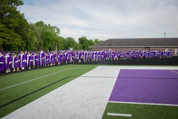 Group of graduates in purple caps and gowns standing on a field during a graduation ceremony