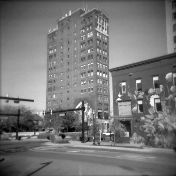 Black and white photo of a tall building next to a smaller building with mural art on the side in an urban street scene.