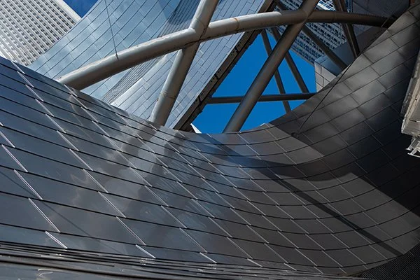 A modern building with reflective glass and metal architectural features, including curved surfaces and supporting beams against a blue sky.