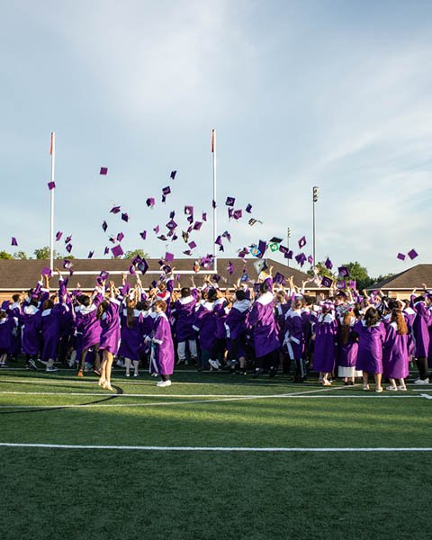 Graduates in purple caps and gowns celebrating on a football field, throwing caps into the air during a graduation ceremony.