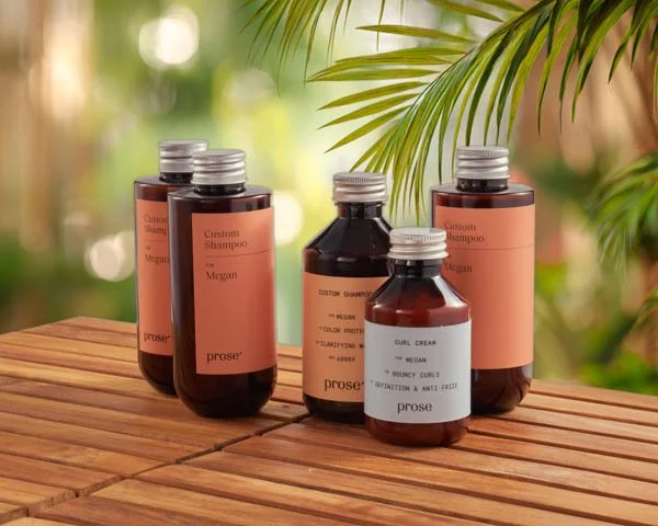 Six brown plastic bottles of shampoo and conditioner on a wooden surface with greenery in the background.