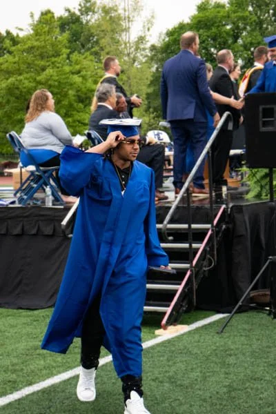 Graduates in blue caps and gowns at an outdoor graduation ceremony on a grassy field, with some seated on chairs and others standing, trees in the background.