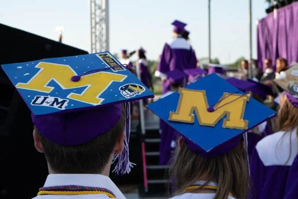 Graduates in caps and gowns at a graduation ceremony with decorated caps featuring the University of Michigan logo and colors.
