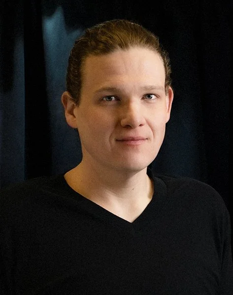 A young person with light skin, brown hair slicked back, wearing a black shirt, standing in front of a dark background.