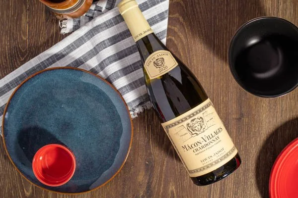 A bottle of wine, a black drinking glass, a blue ceramic plate, and a red shot glass on a wooden table with a gray and white striped napkin.