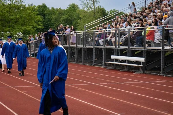 Graduates in blue caps and gowns walking on a track during a graduation ceremony, with an audience seated in bleachers in the background.